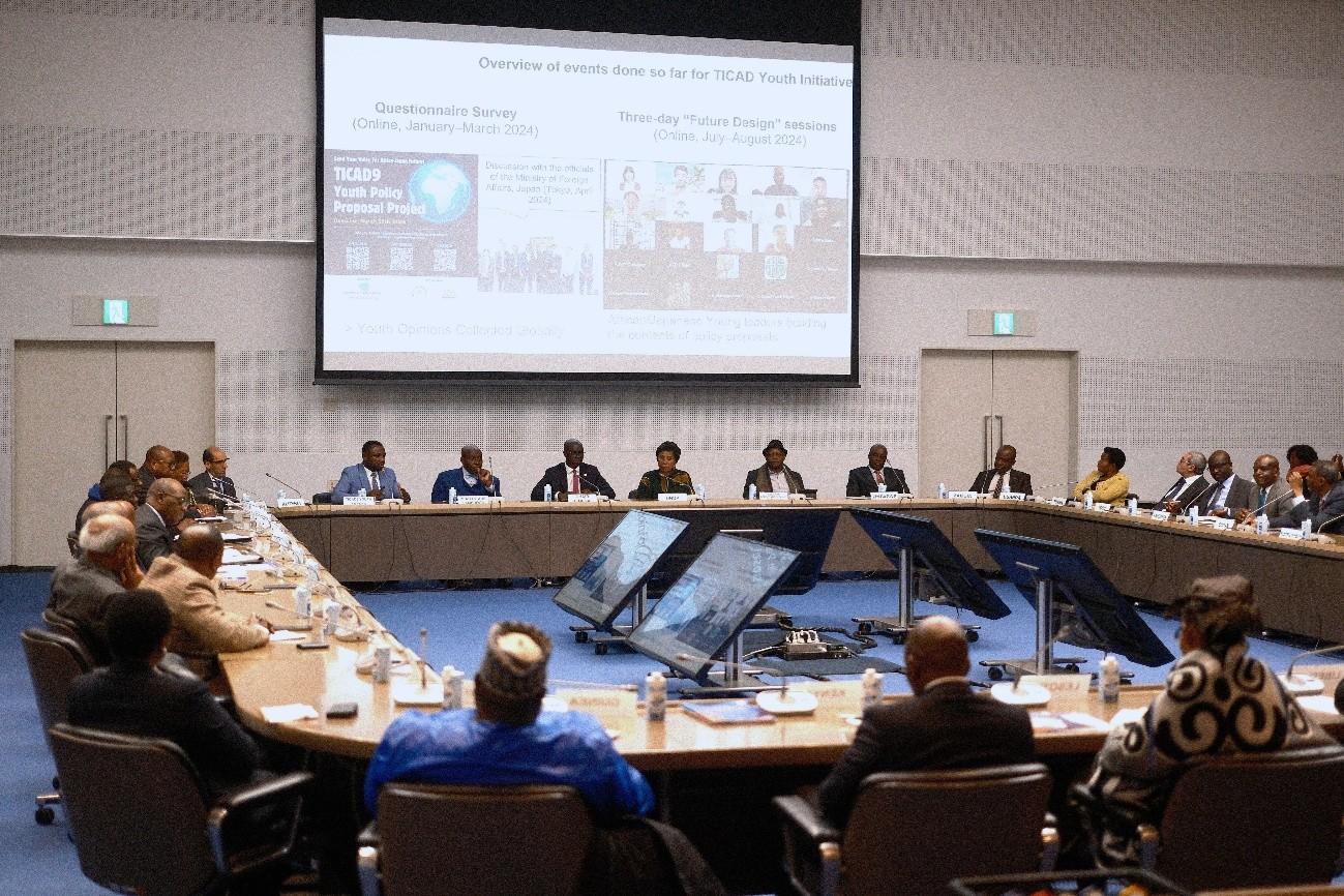 A conference room filled with people seated around a large table, with a presentation on a screen.
