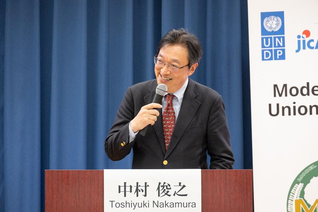A man in a suit speaks at a podium with a microphone, smiling.