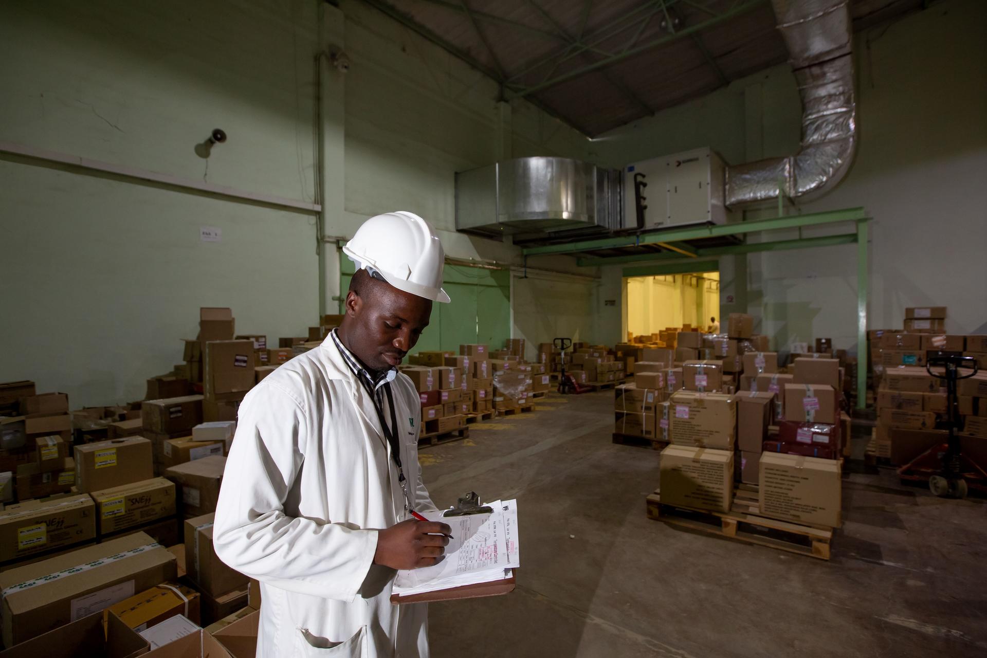 Warehouse worker in a white coat and hard hat reviewing notes among boxes.