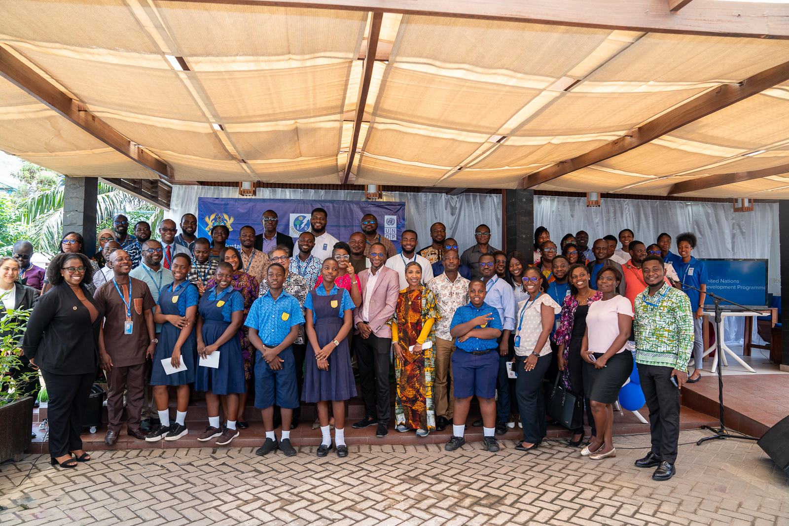 A diverse group of adults and schoolchildren poses together under a shaded area.