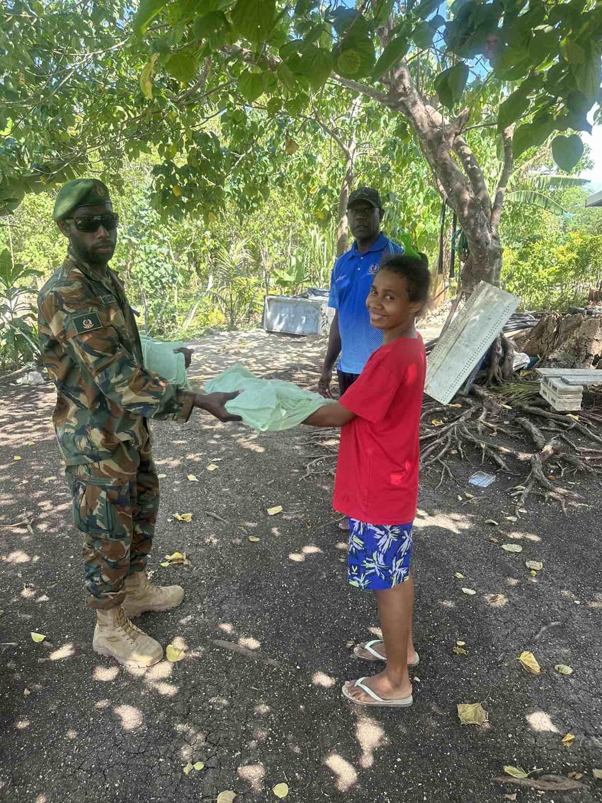A child in a red shirt receives material from a soldier in camouflage under a tree.