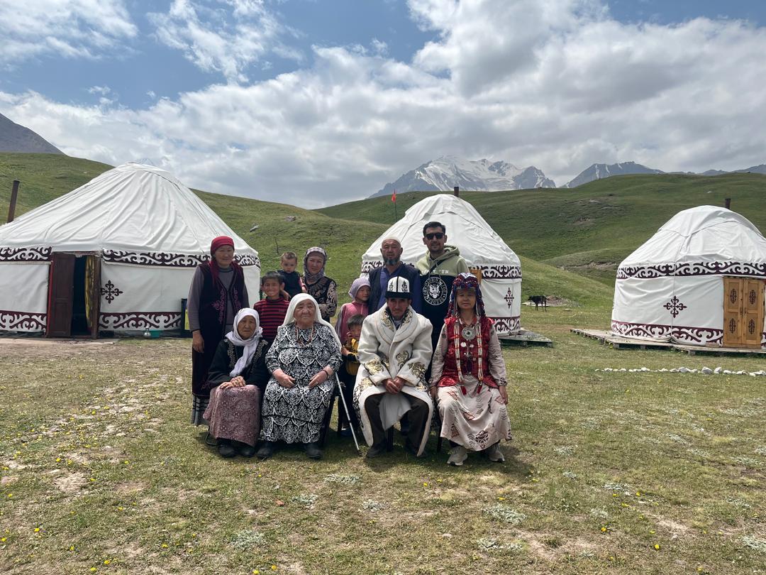 A group of people dressed in traditional attire stands in front of yurts on a grassy landscape.