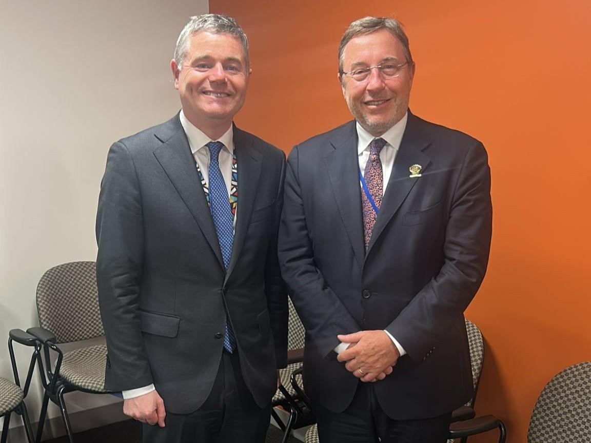Two men in business suits smiling against an orange wall, chairs in the background.