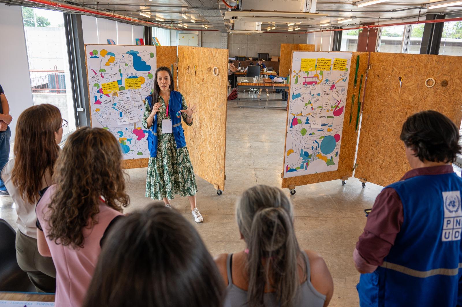 A presenter stands in front of a group, gesturing towards colorful visual displays on boards.