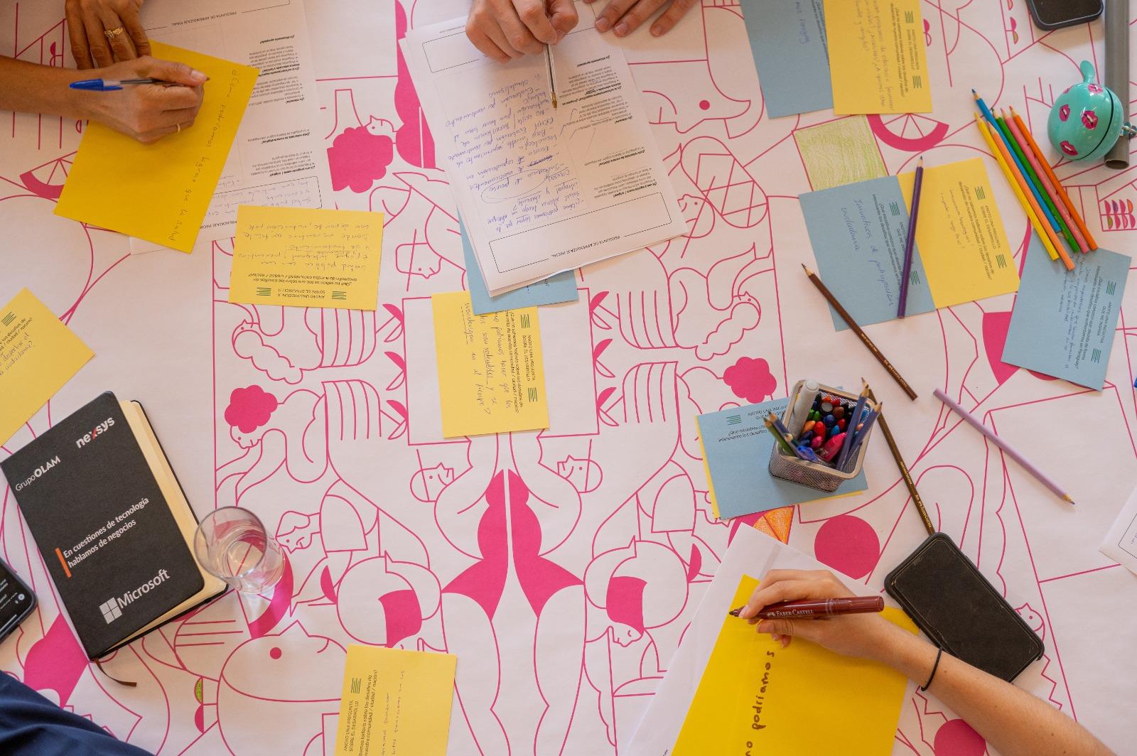 A group of hands writing on colorful sticky notes on a patterned tablecloth.