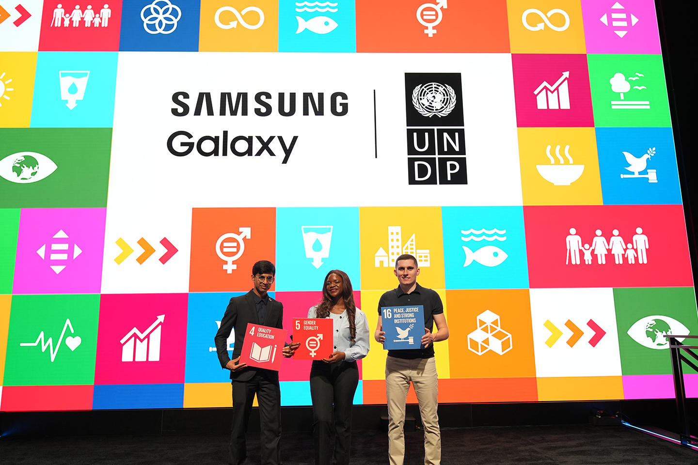 Three people holding signs stand in front of a colorful backdrop featuring logos and icons.