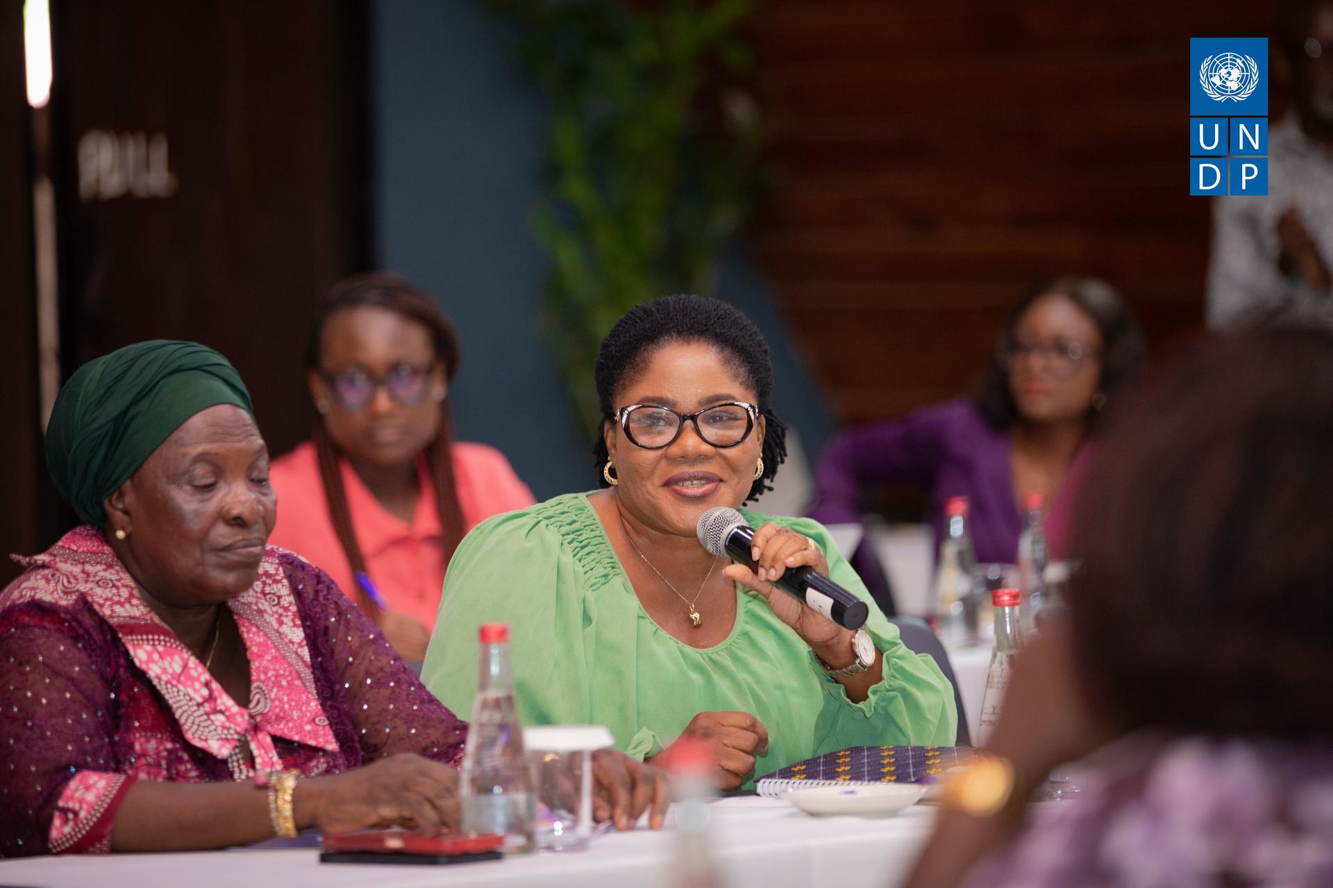 A woman in a green outfit speaks at a round table with attentive listeners in the background.