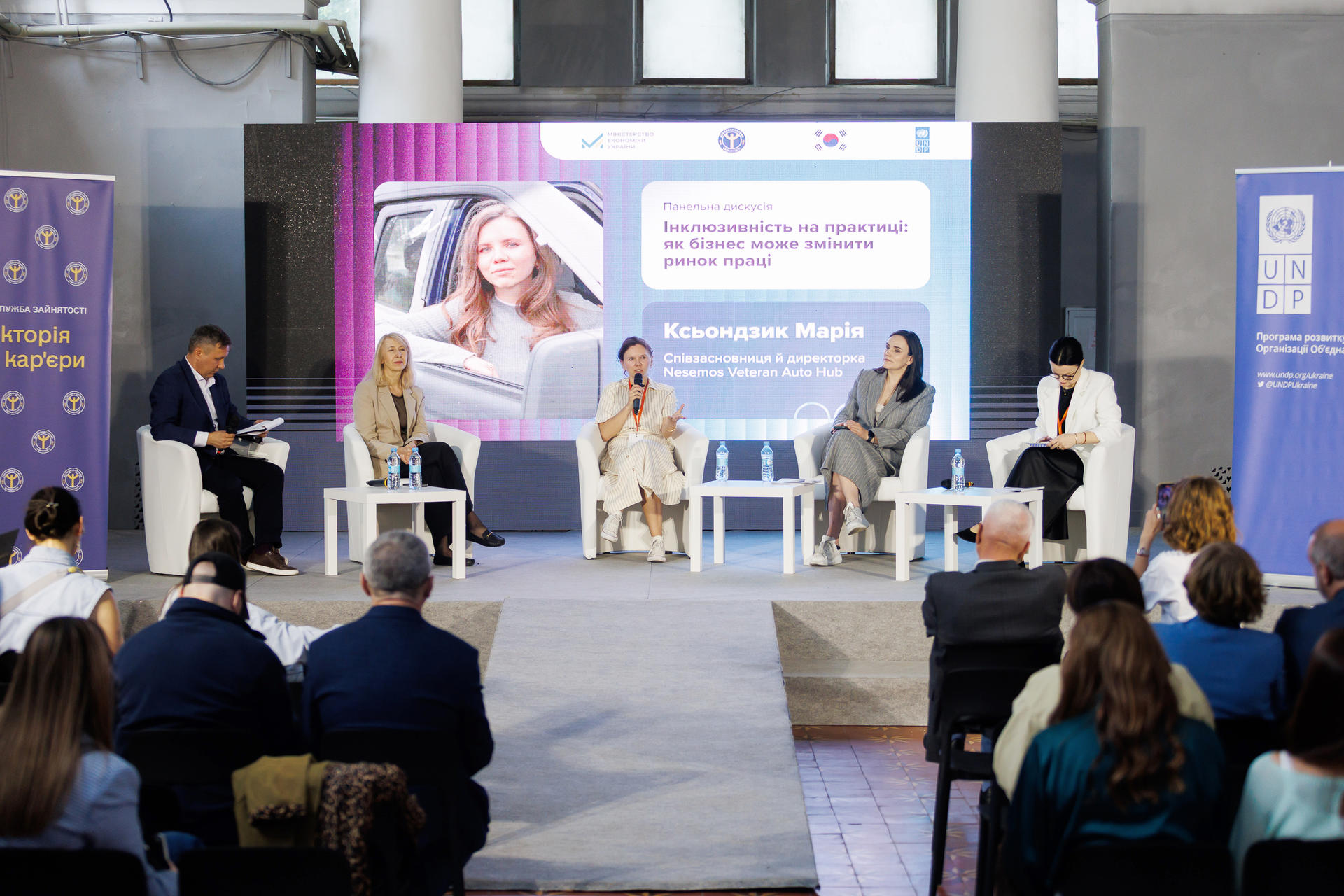 Panel discussion with five speakers on stage, audience in foreground, large screen behind.
