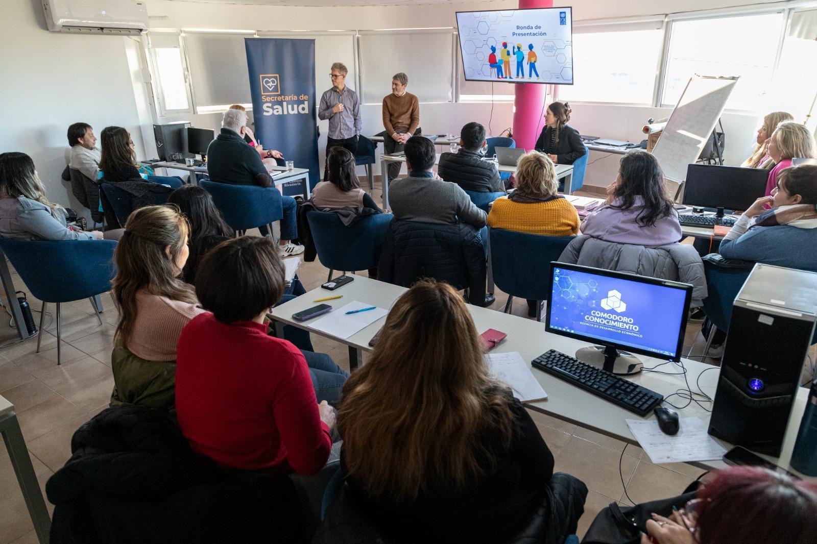 Un grupo de personas mirando una presentación en una sala luminosa con computadoras y una pantalla.