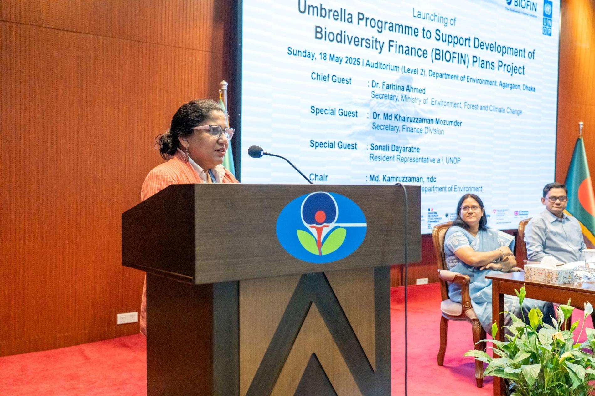 A woman speaks at a podium during a conference on biodiversity finance, with attendees seated nearby.