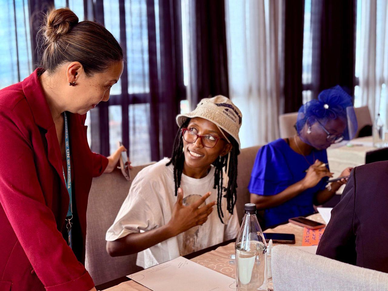 A woman in a beige hat smiles while speaking to another woman in a red blazer at a conference table.