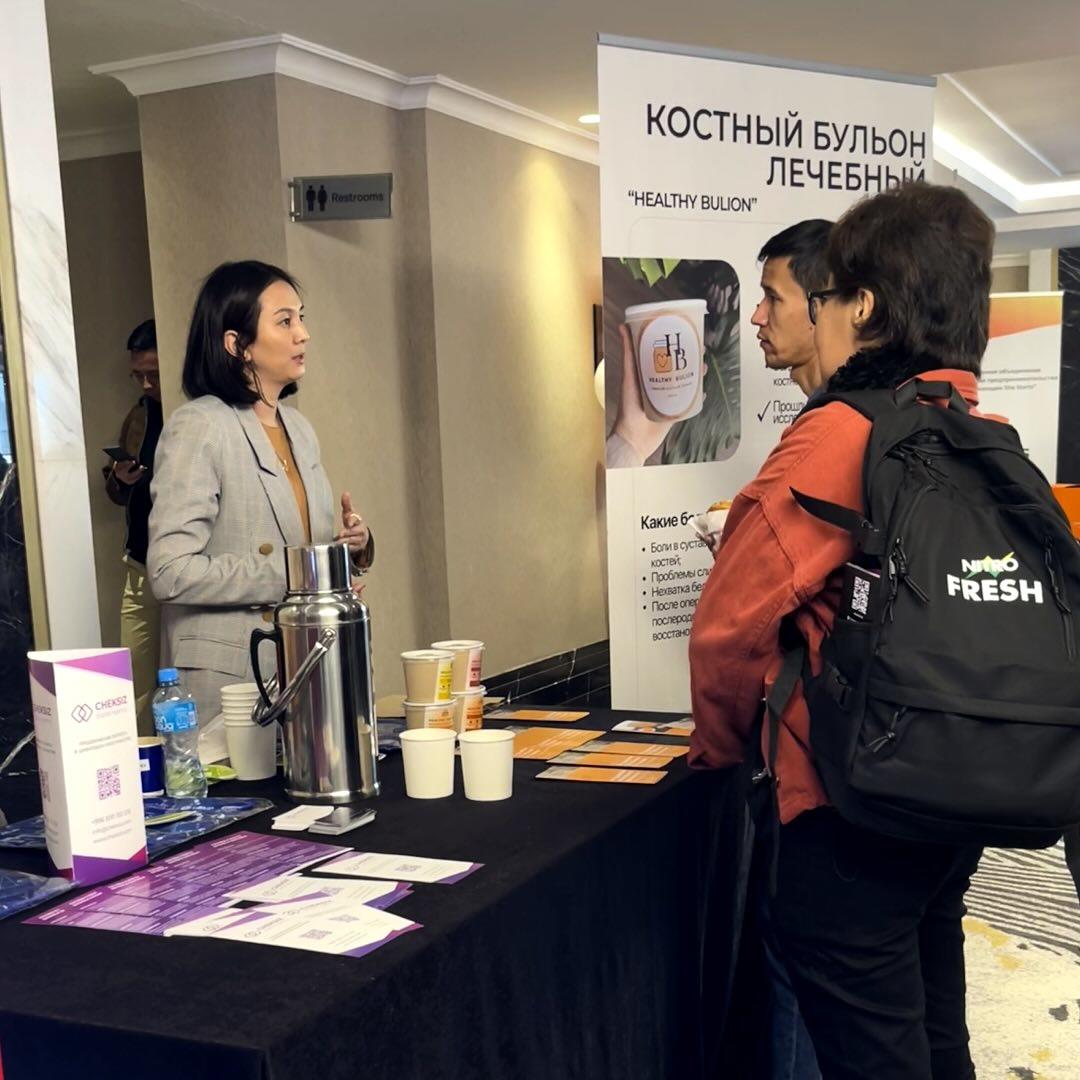 A woman in a gray blazer talks to two attendees at a conference booth featuring health products.