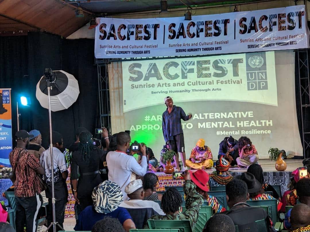 A vibrant festival scene with people gathered, a speaker on stage, and colorful decorations.