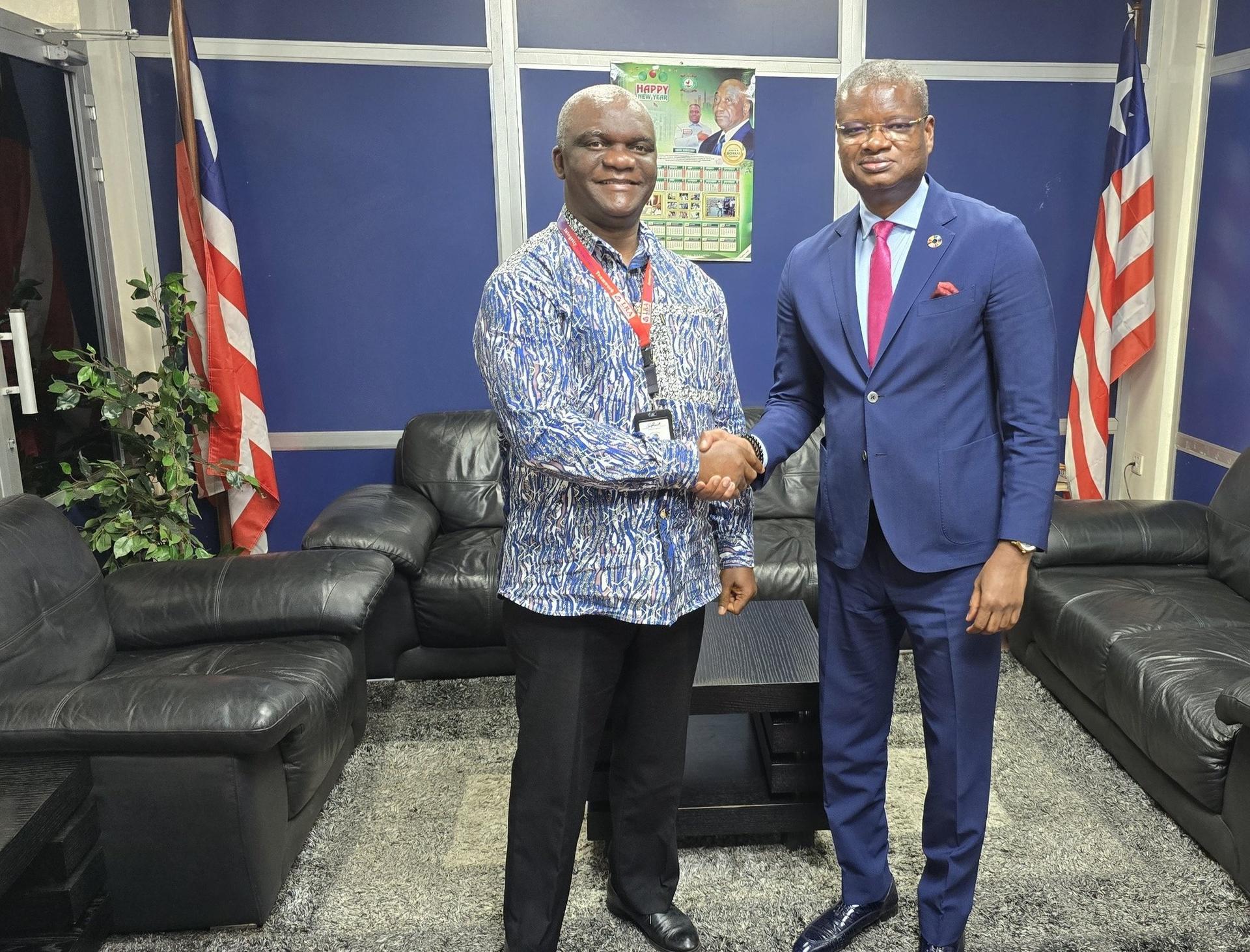 Two men shake hands in a modern office setting, with couches and flags in the background.