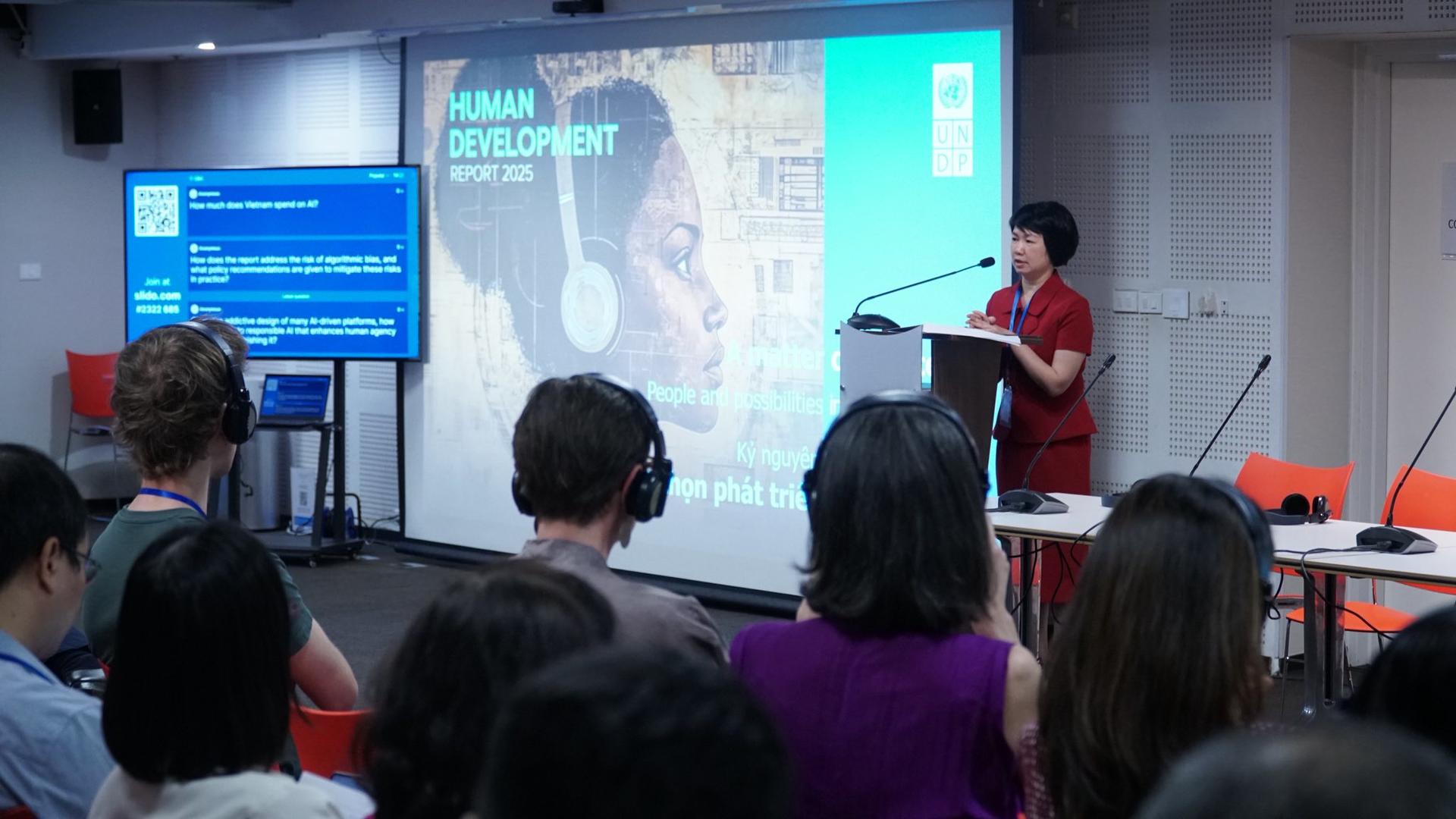 A speaker presents at a conference, with an audience and a large projected slide behind.