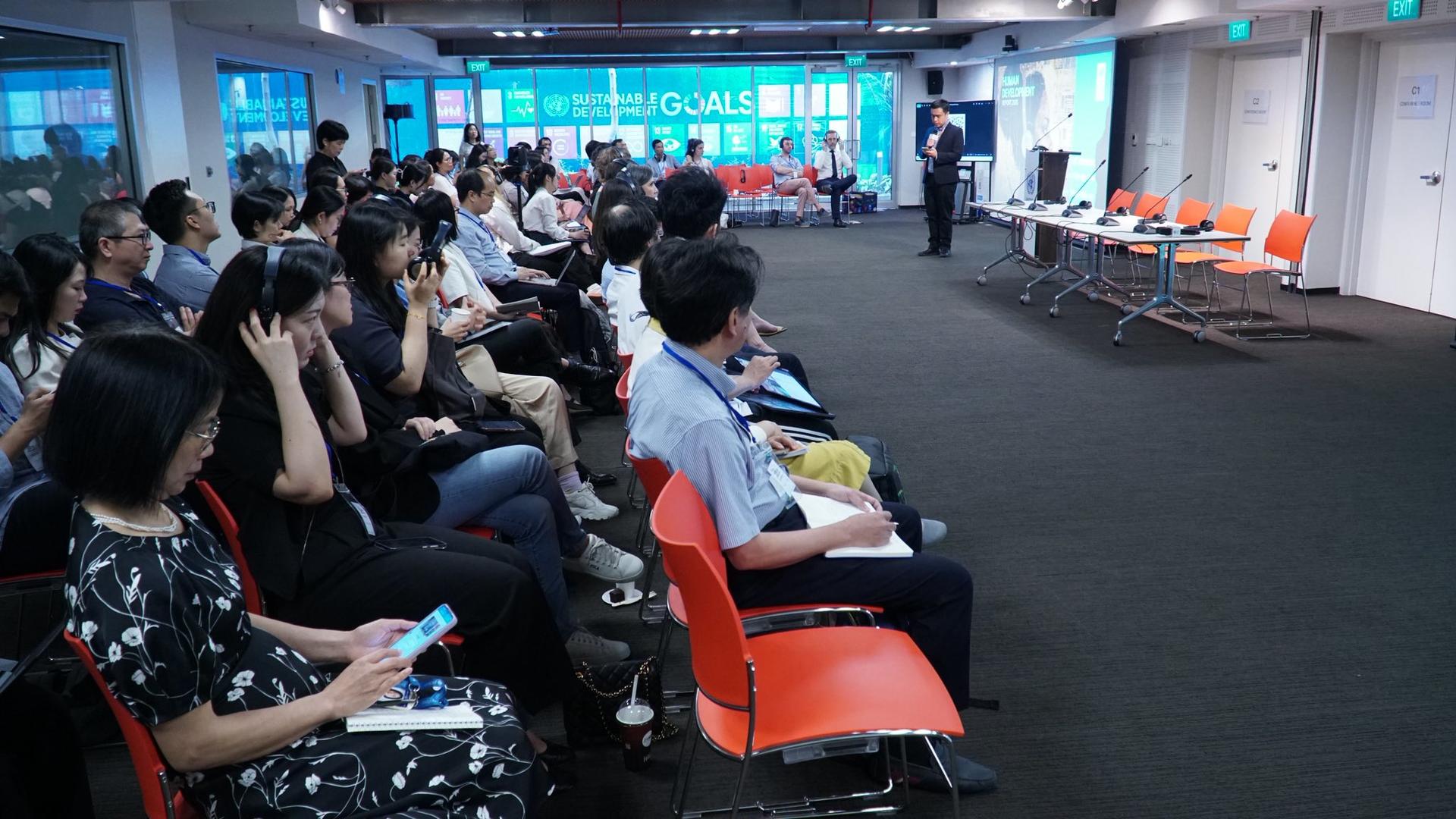 Audience in a modern conference room, facing a speaker at the front.