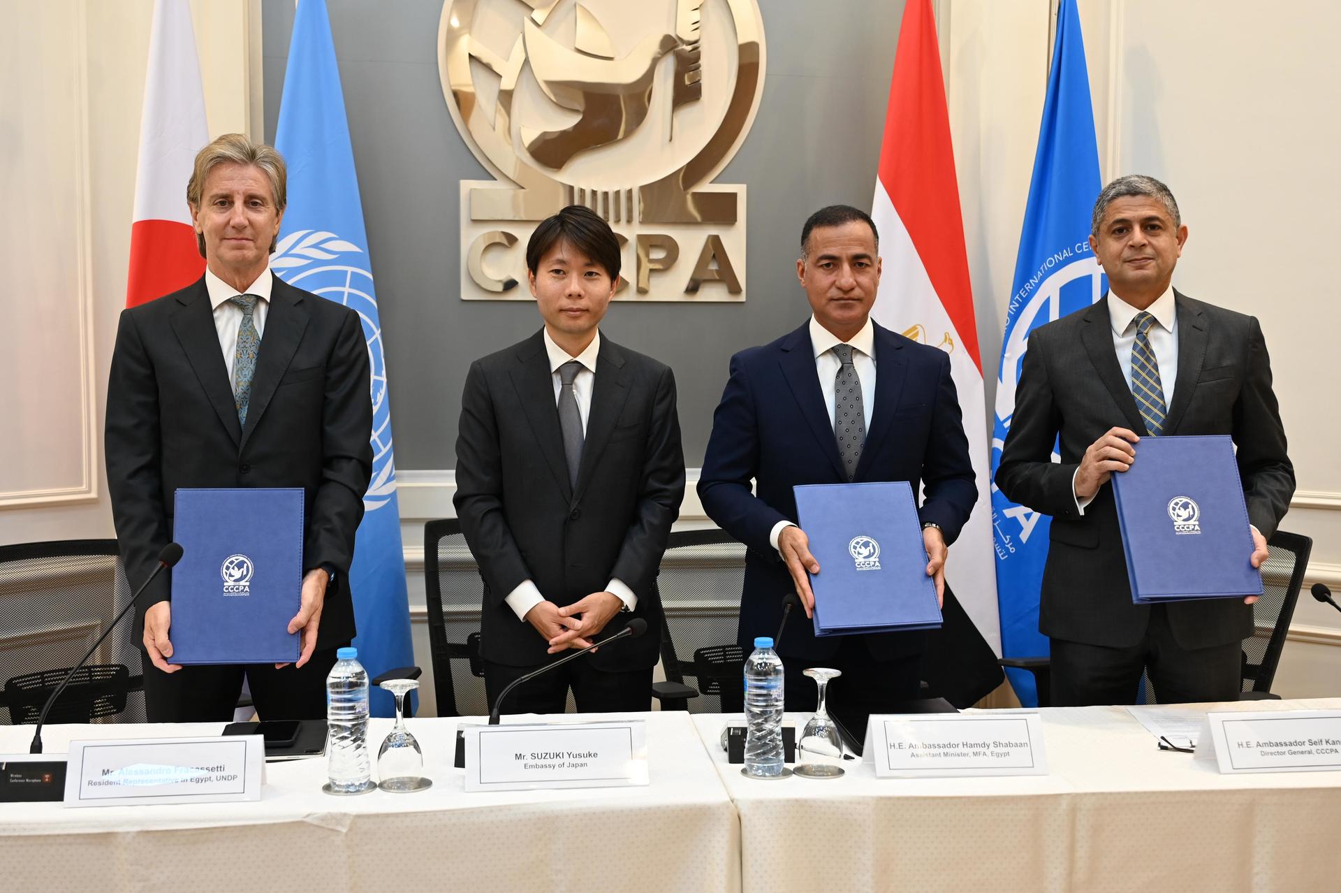 Four men holding blue documents stand at a table with flags in the background.