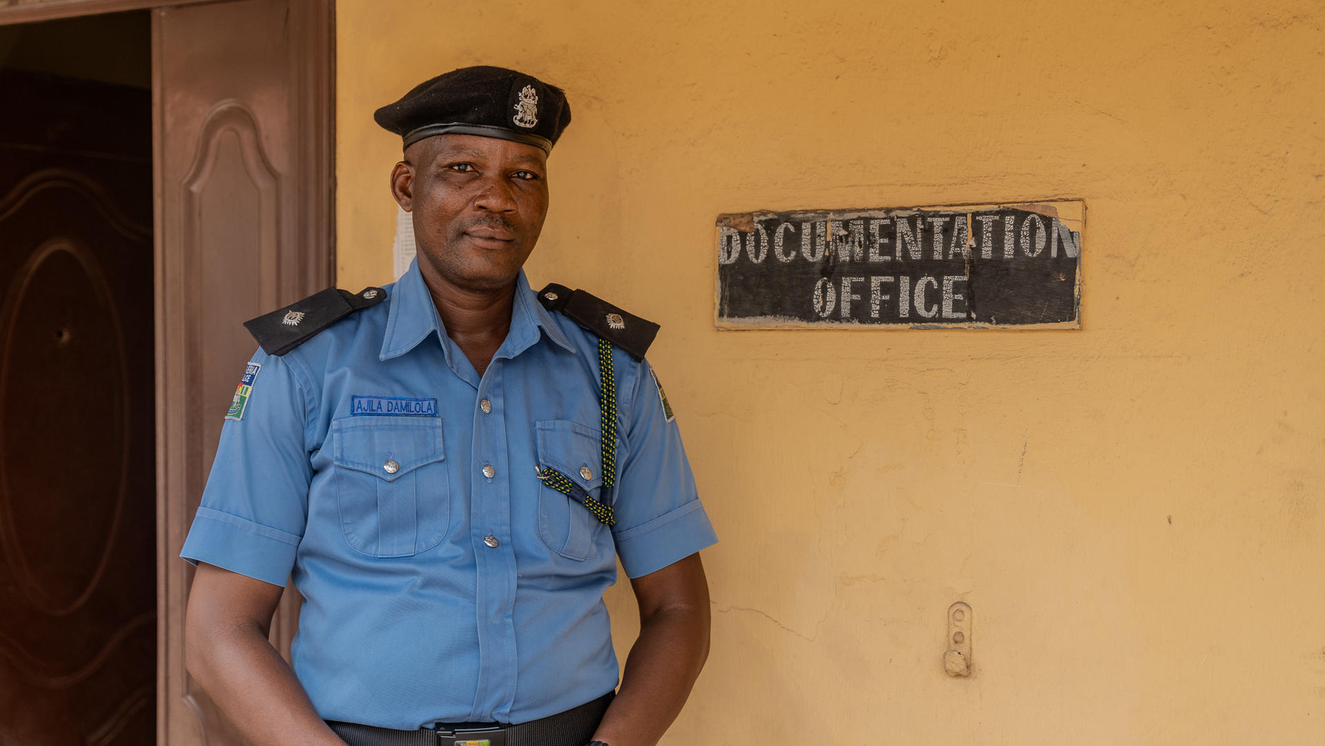 A police officer stands smiling next to a "Documentation Office" sign on a yellow wall.