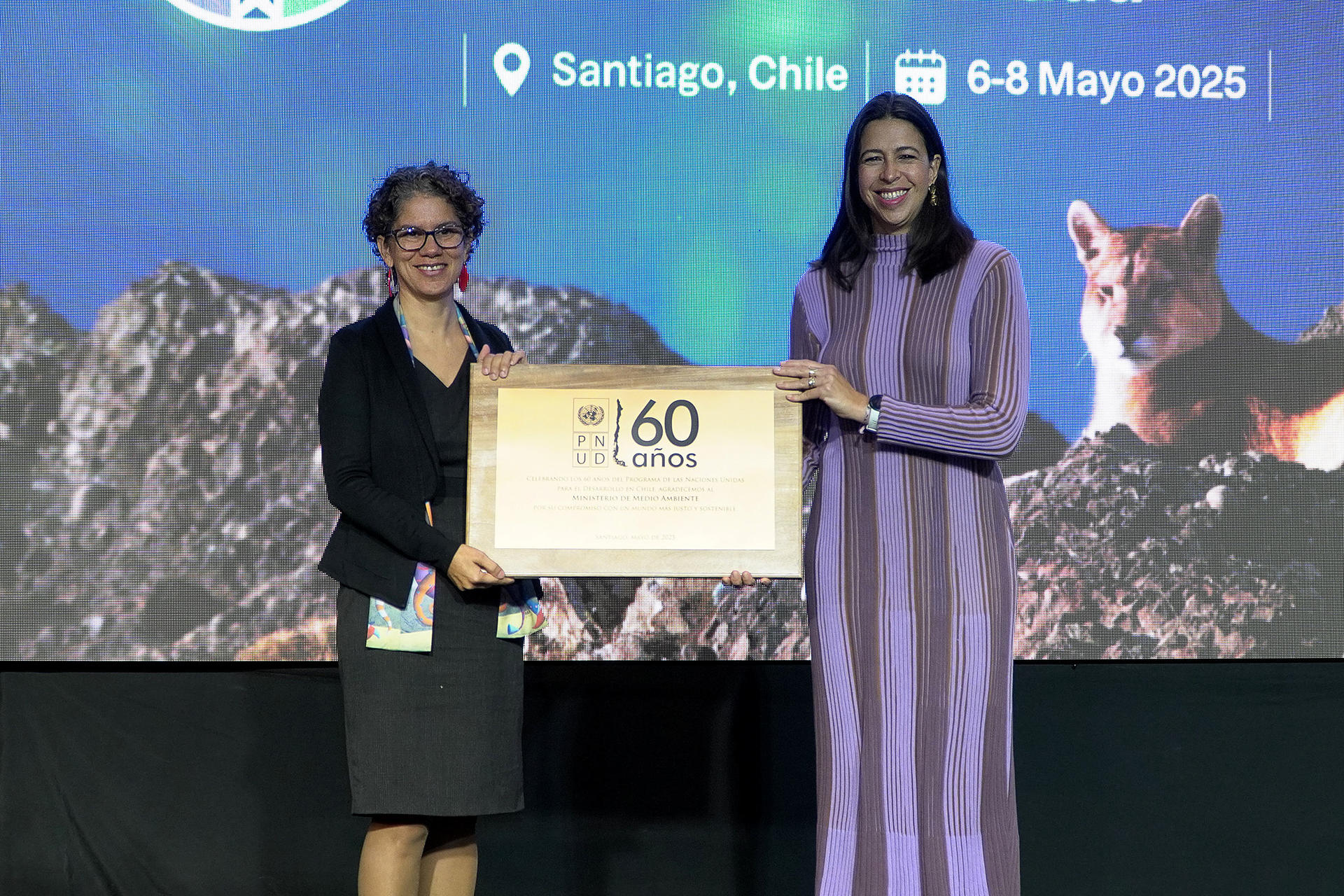 Two women are on stage, one handing a wooden plaque to the other, with a scenic backdrop.
