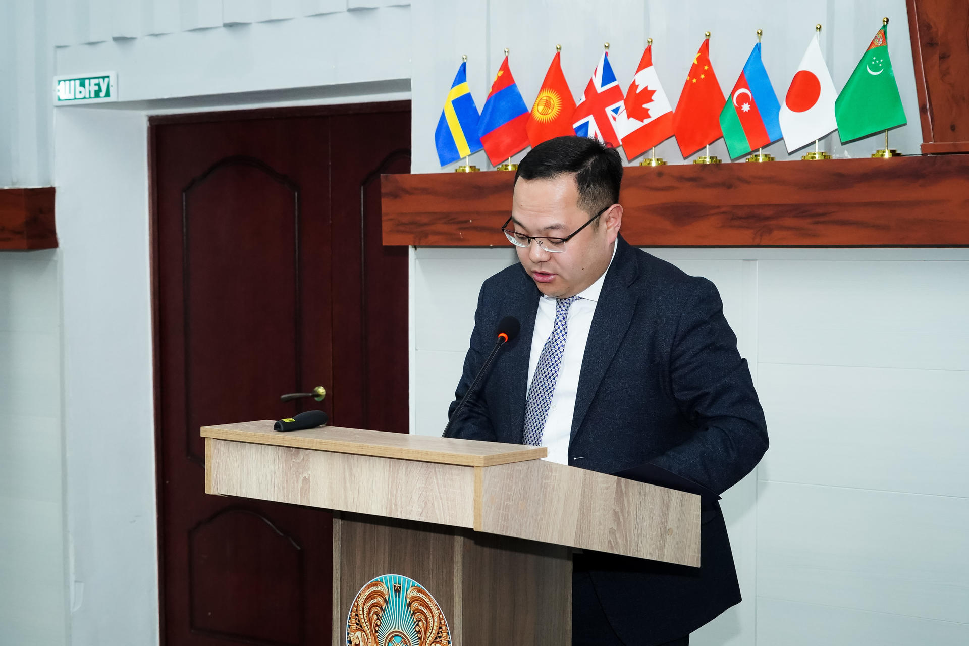 A man in a suit delivers a speech at a podium, with flags in the background.