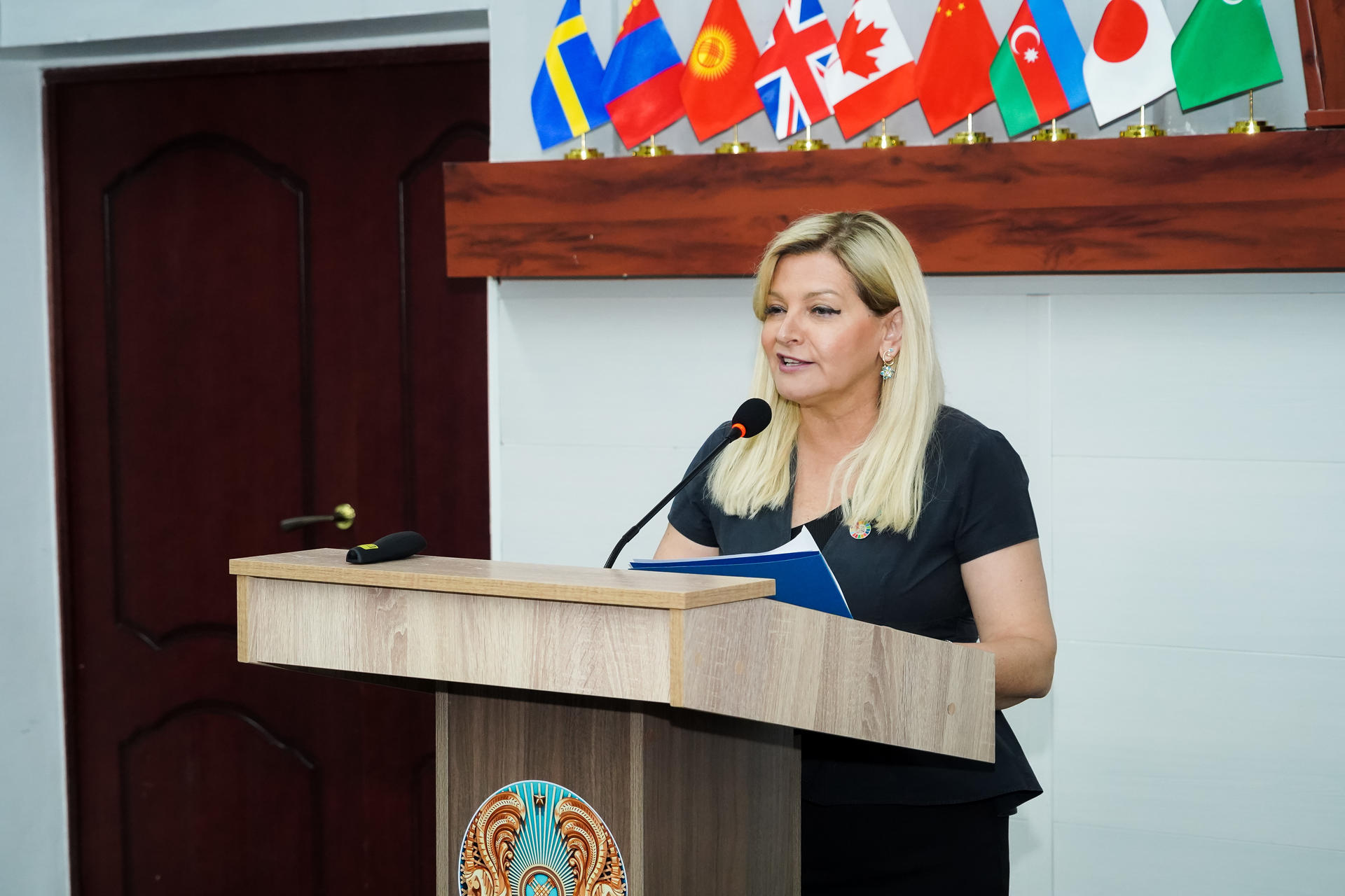 A woman speaks at a podium with various national flags displayed behind her.