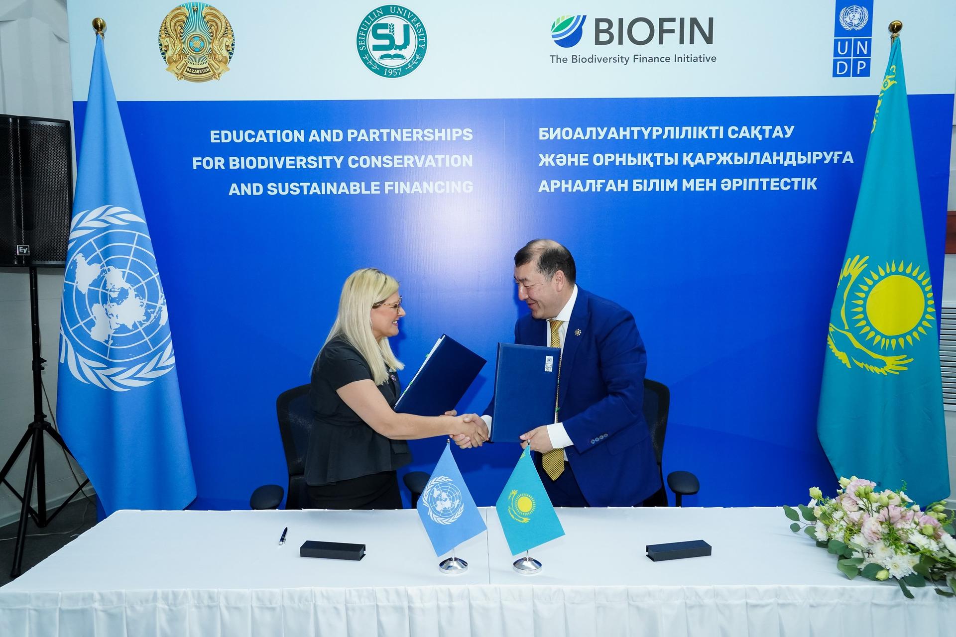 Two individuals shaking hands while exchanging signed documents at a table, with flags in the background.