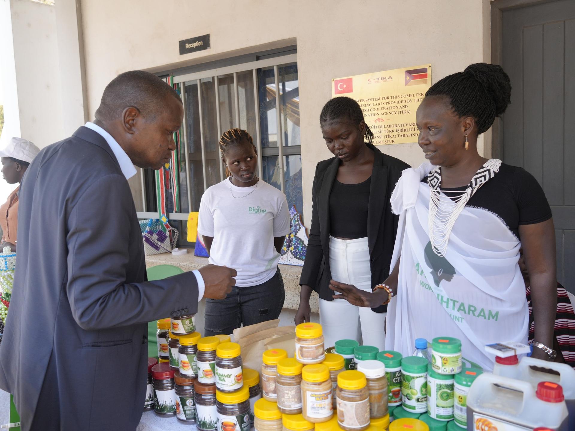 A man converses with three women at a market stall filled with various jars and containers.