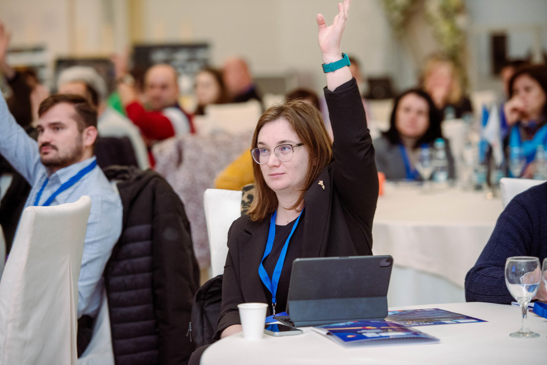 A woman with glasses raises her hand in a conference setting, surrounded by attendees.