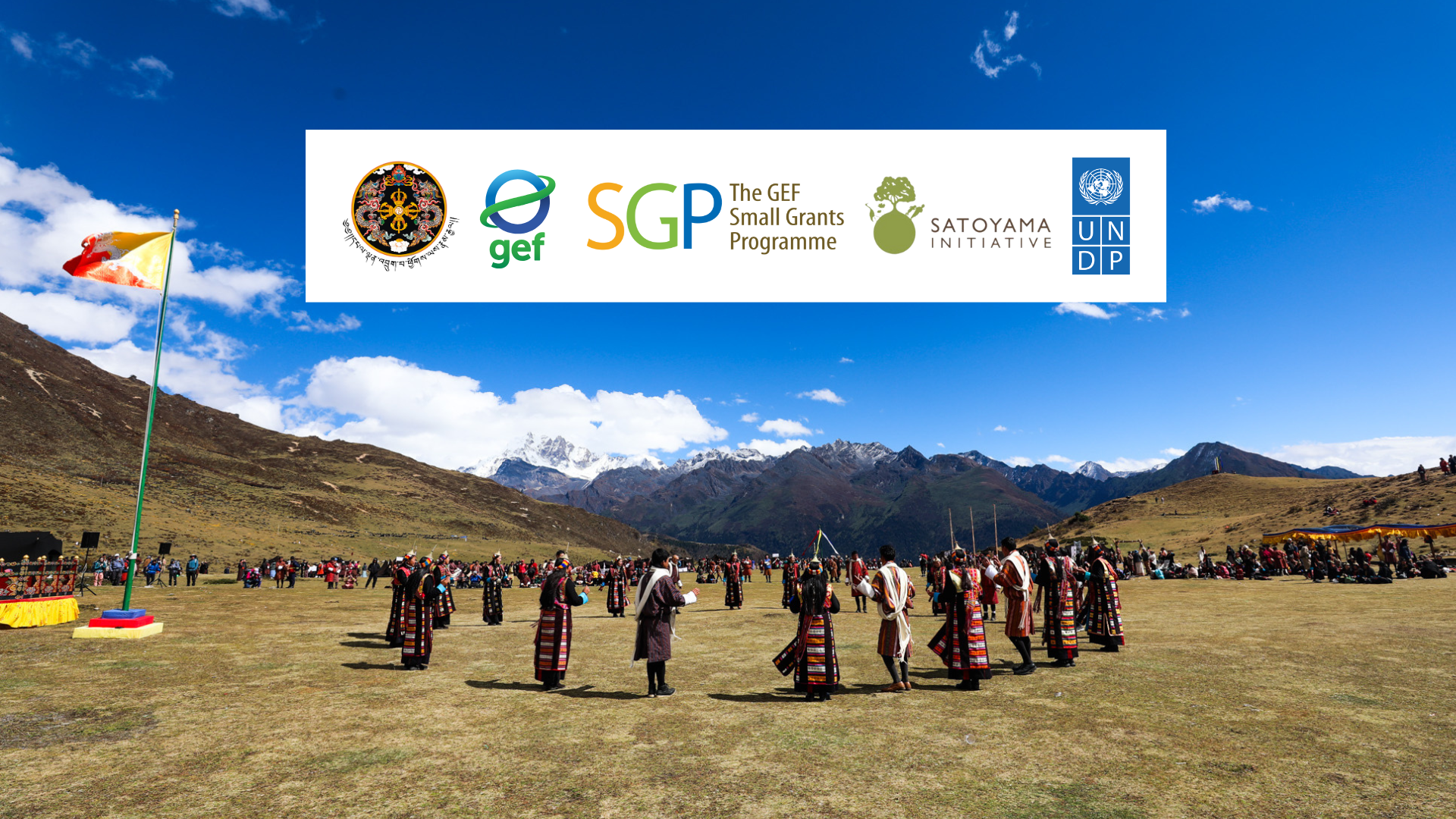 People in traditional clothing dance on a grassy field with mountains in the background.