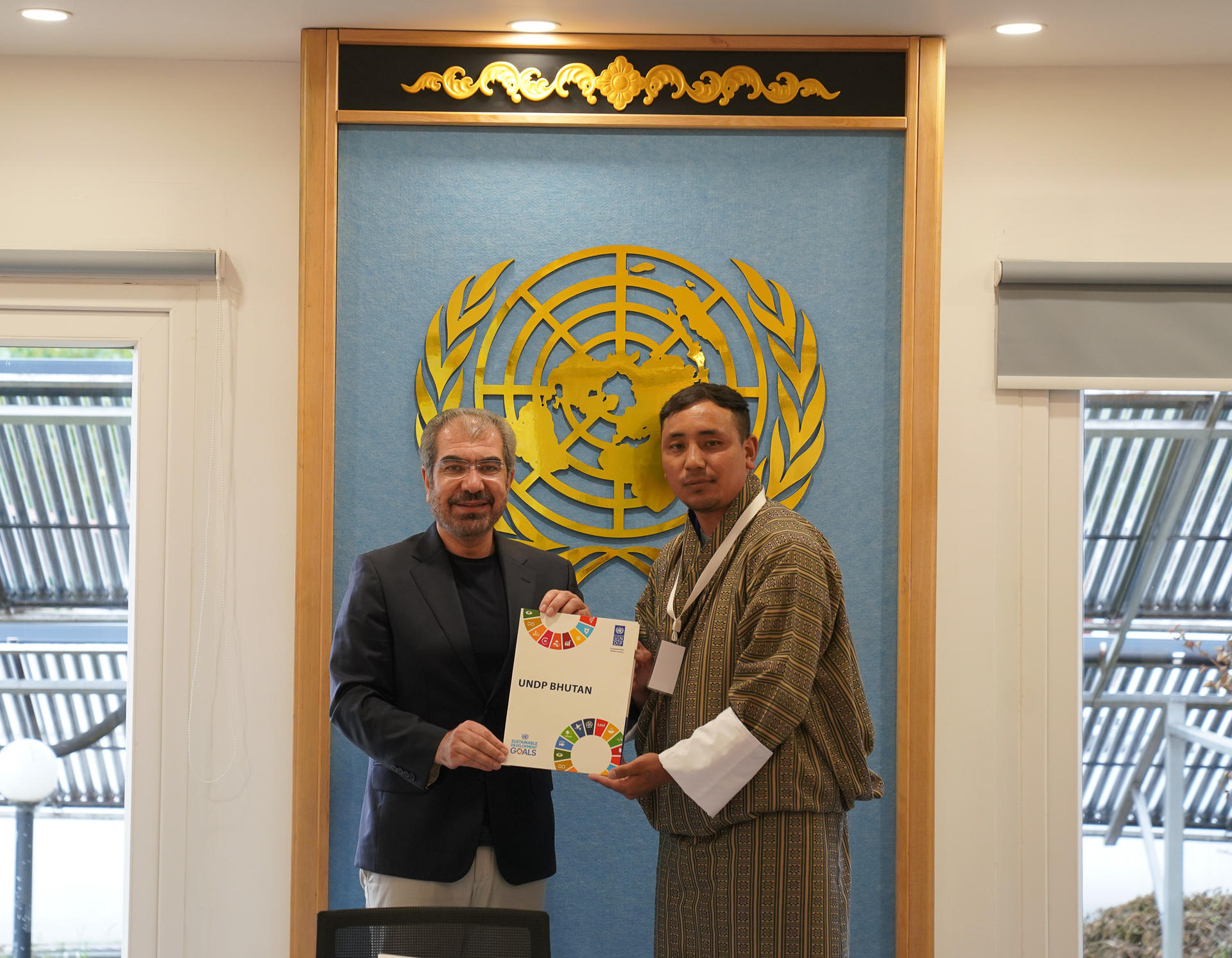 Two men stand smiling while holding a certificate in front of a UN emblem backdrop.