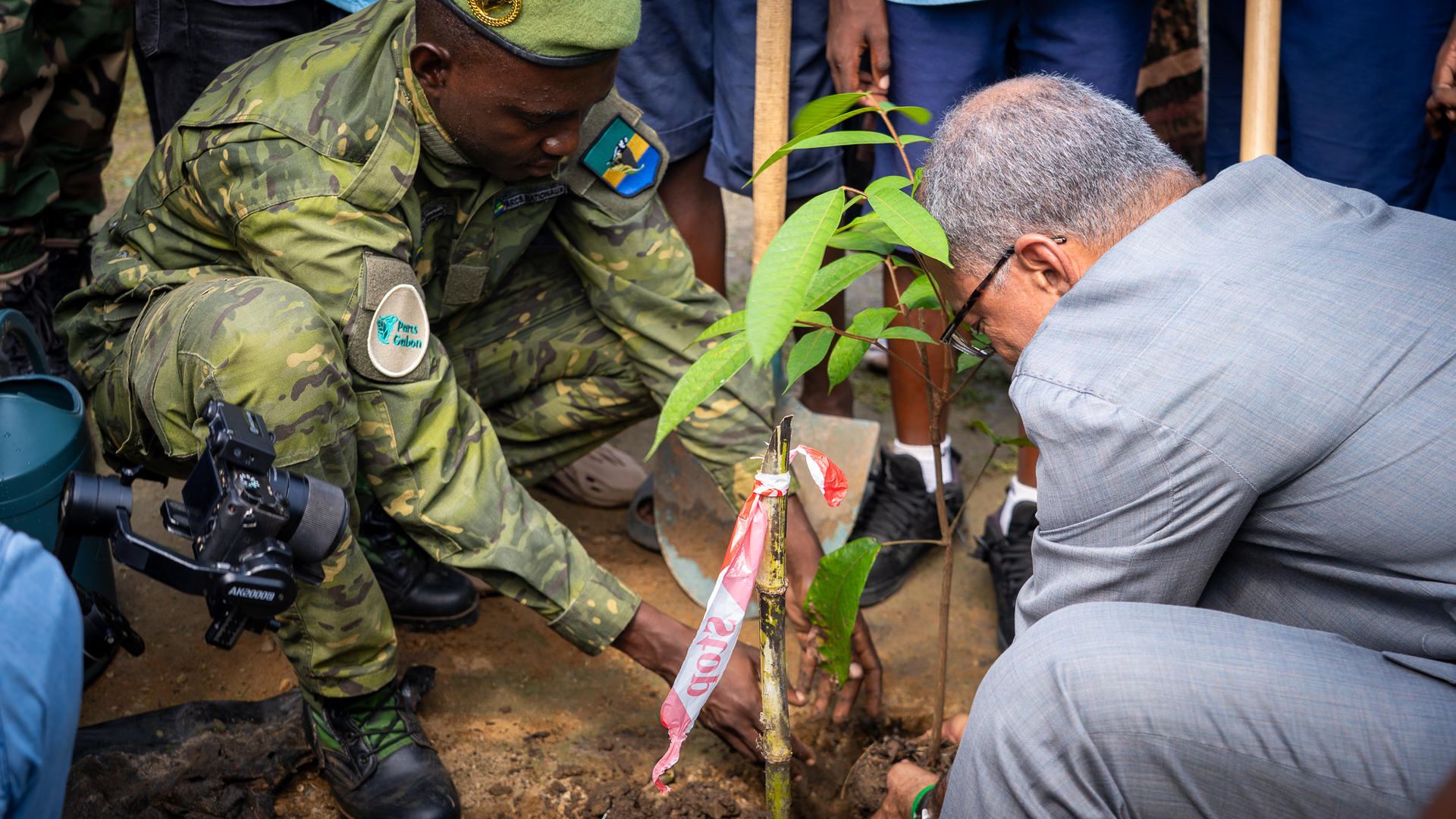 A soldier and a man in a suit plant a young tree surrounded by onlookers.
