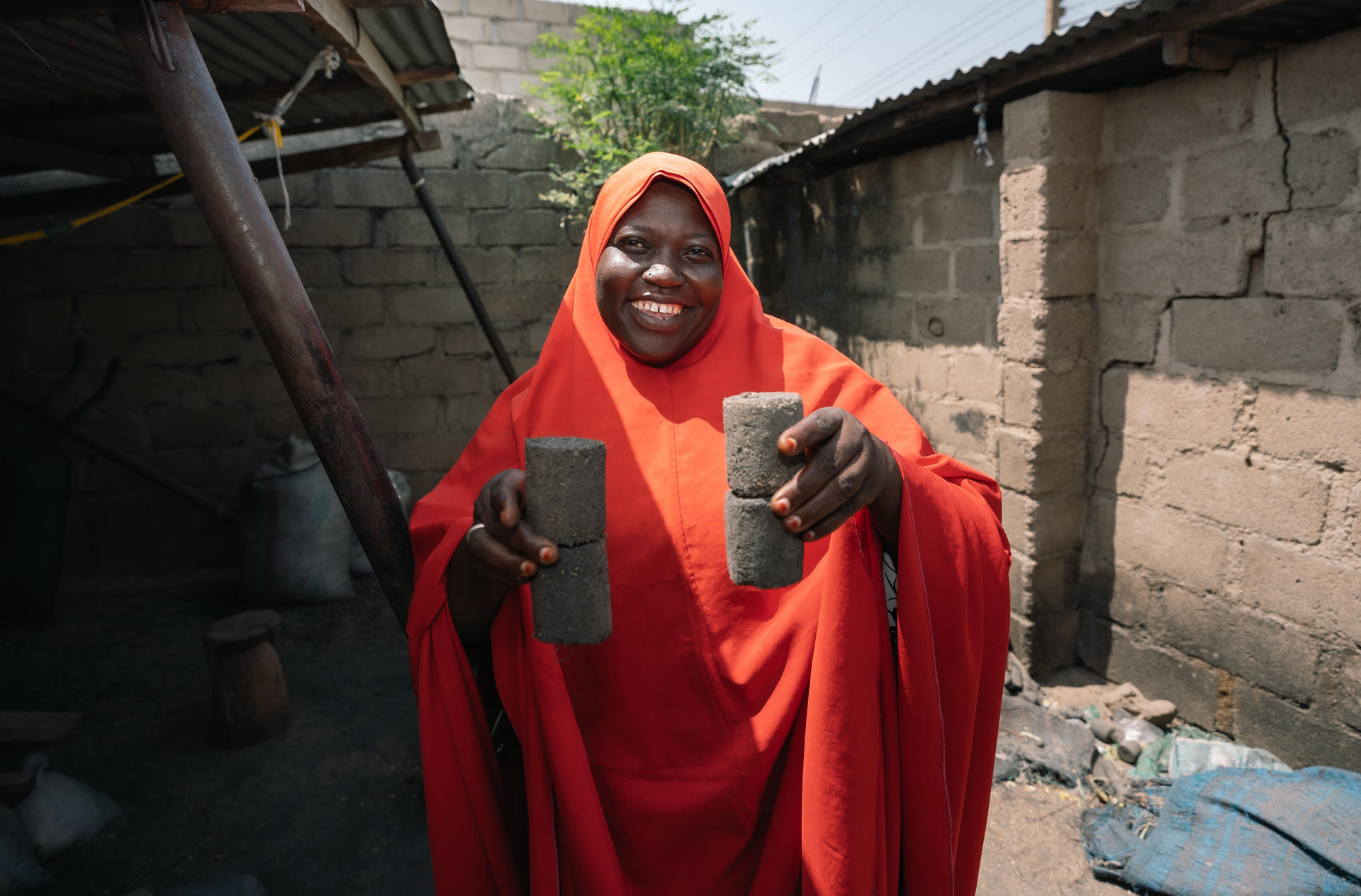 A smiling woman in a red cloak holding two cylindrical stones in an outdoor setting.