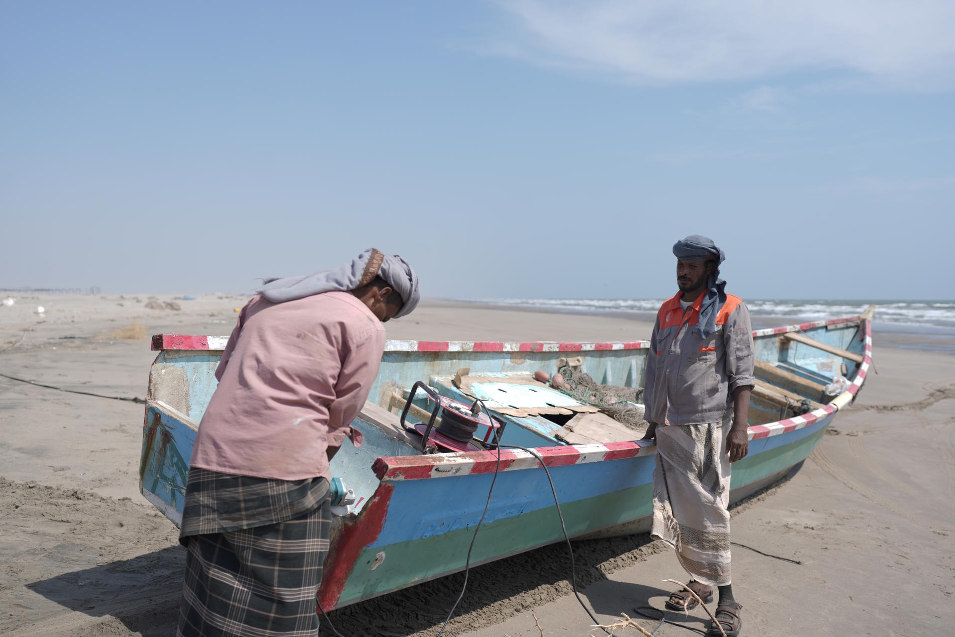 Two men working on a colorful fishing boat on a sandy beach under a clear blue sky.