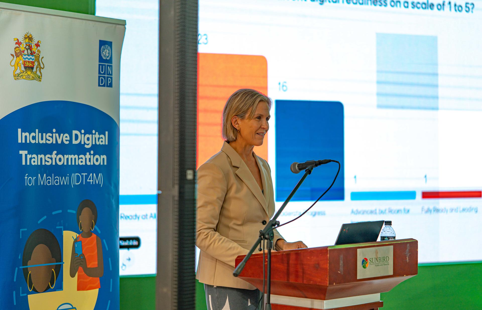 A woman in a blazer stands at a podium, speaking with a digital presentation behind her.