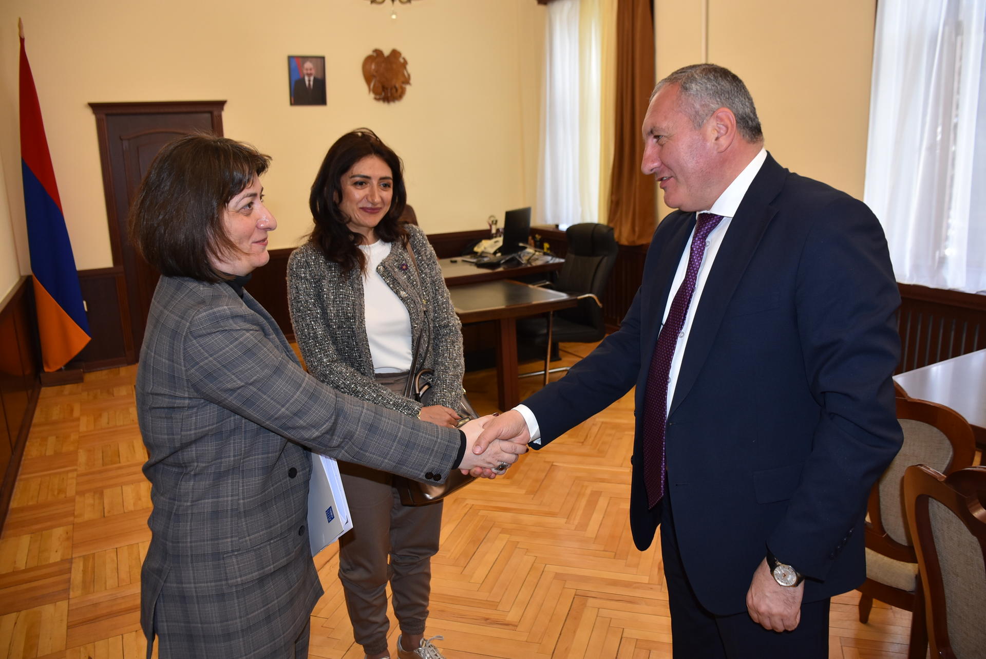 A man and a woman shake hands in an office, with a second woman observing.