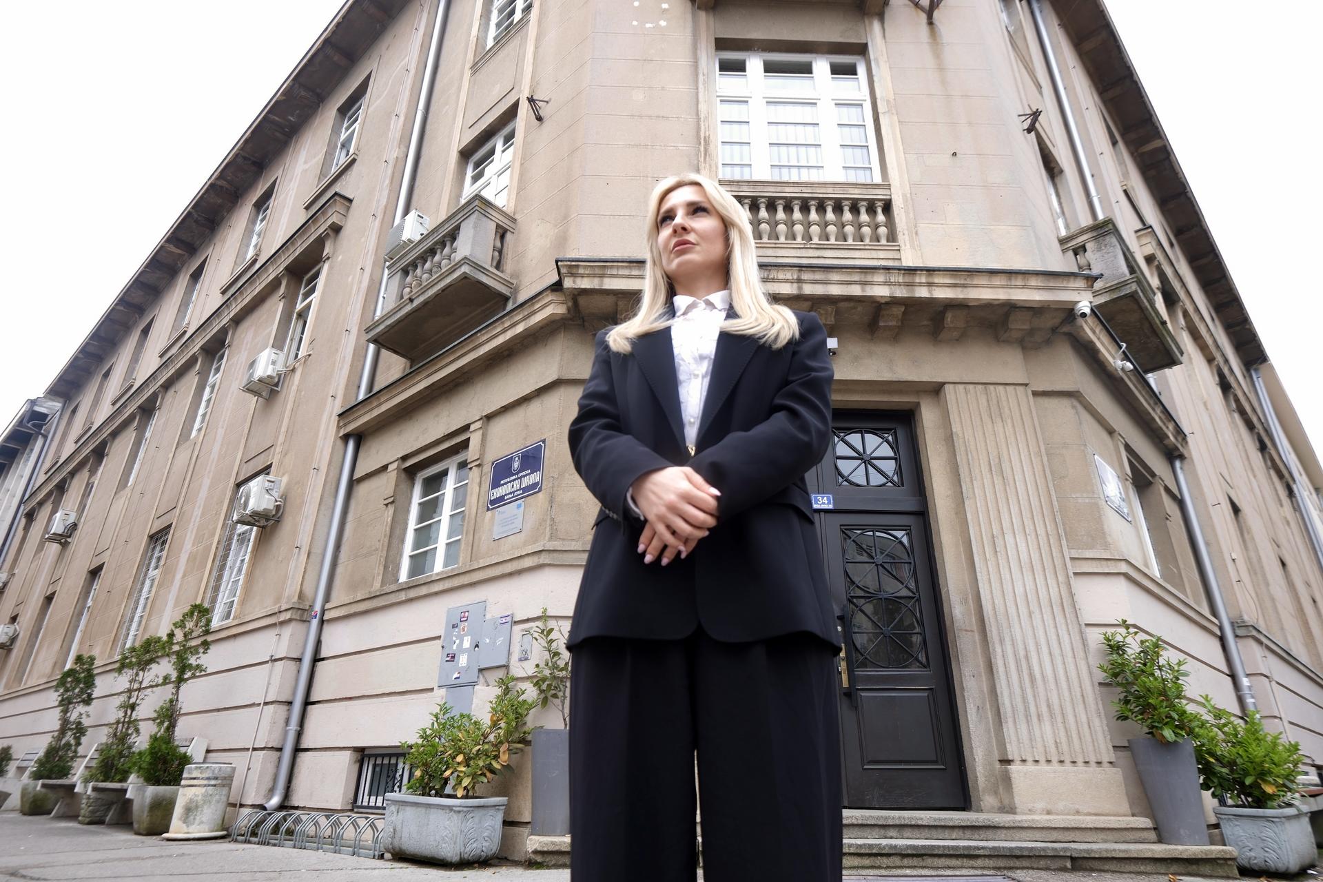 A woman in a black suit stands confidently outside a building, with potted plants nearby.