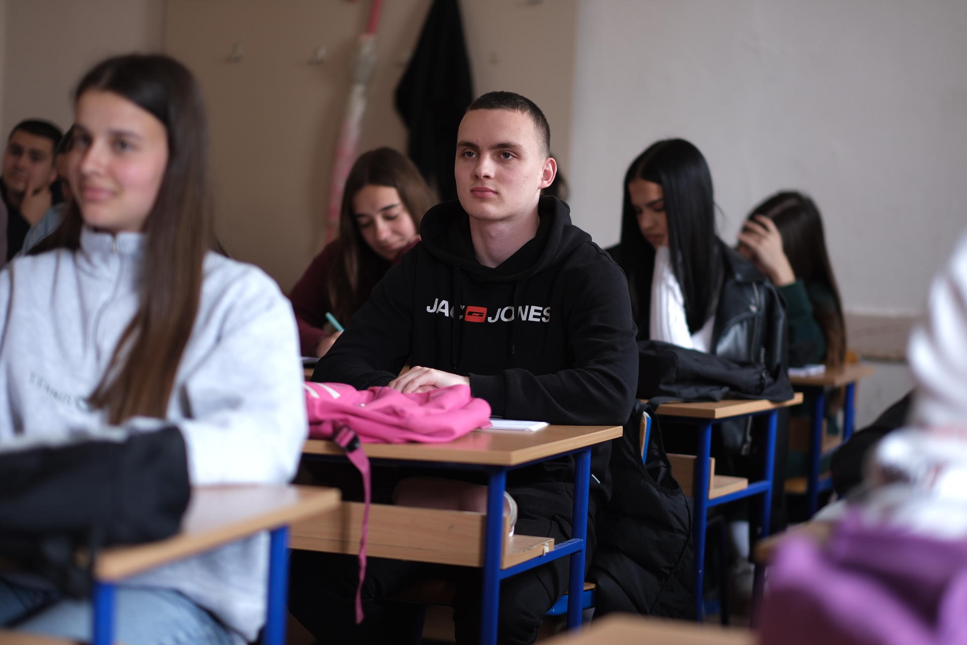 A classroom scene with students seated, one boy in a black hoodie focused, others engaged.