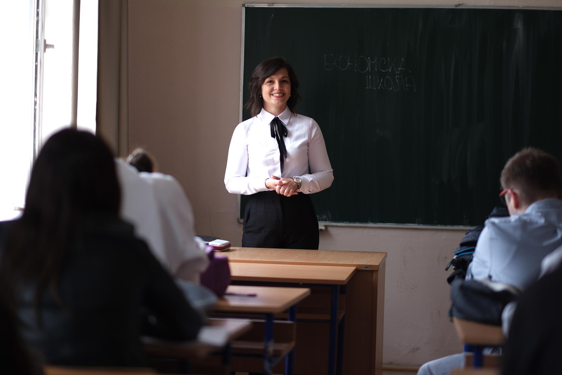 A teacher standing in front of a chalkboard, with students visible at their desks.
