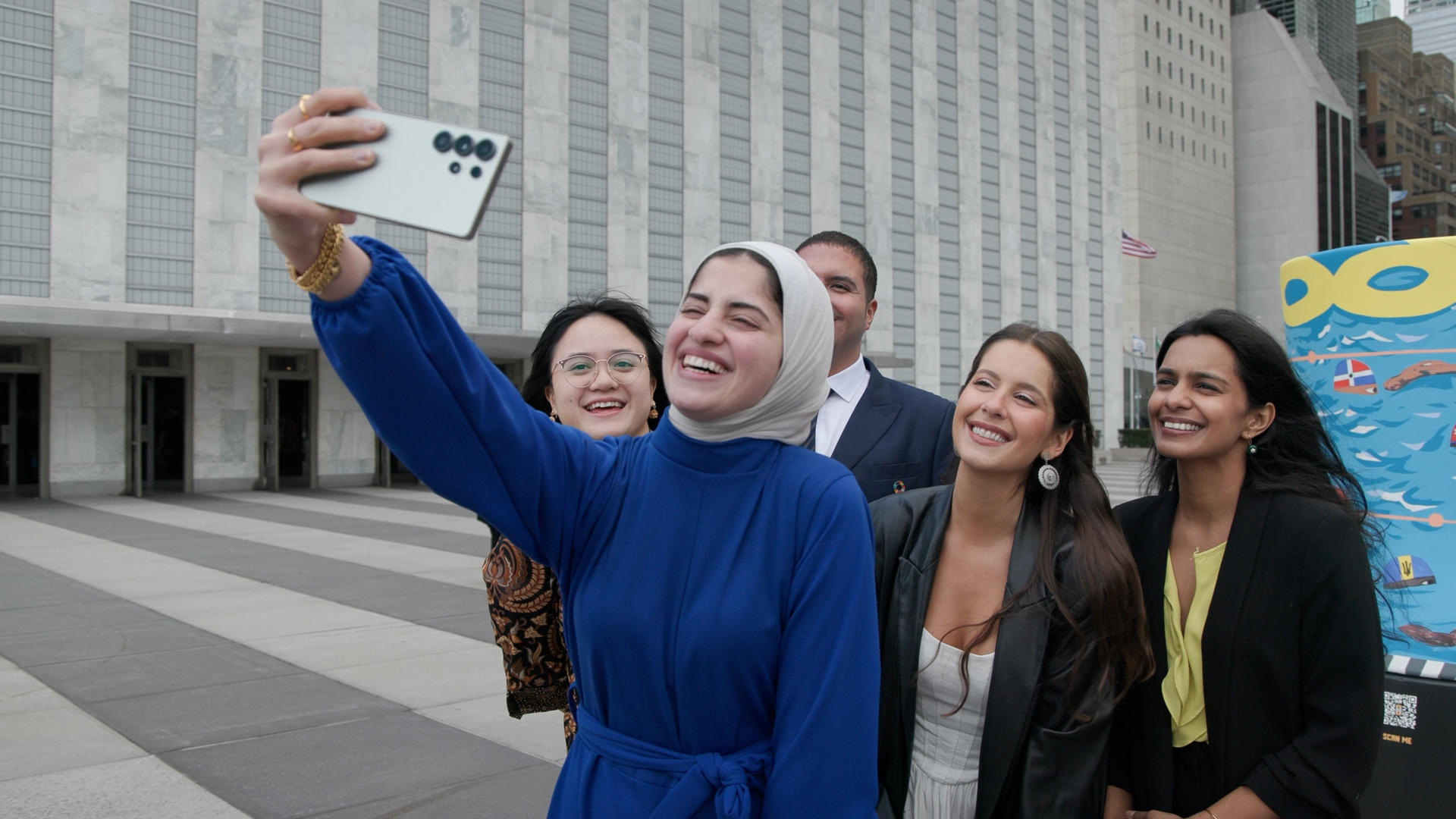 A group of five people taking a selfie outside a modern building, smiling happily.