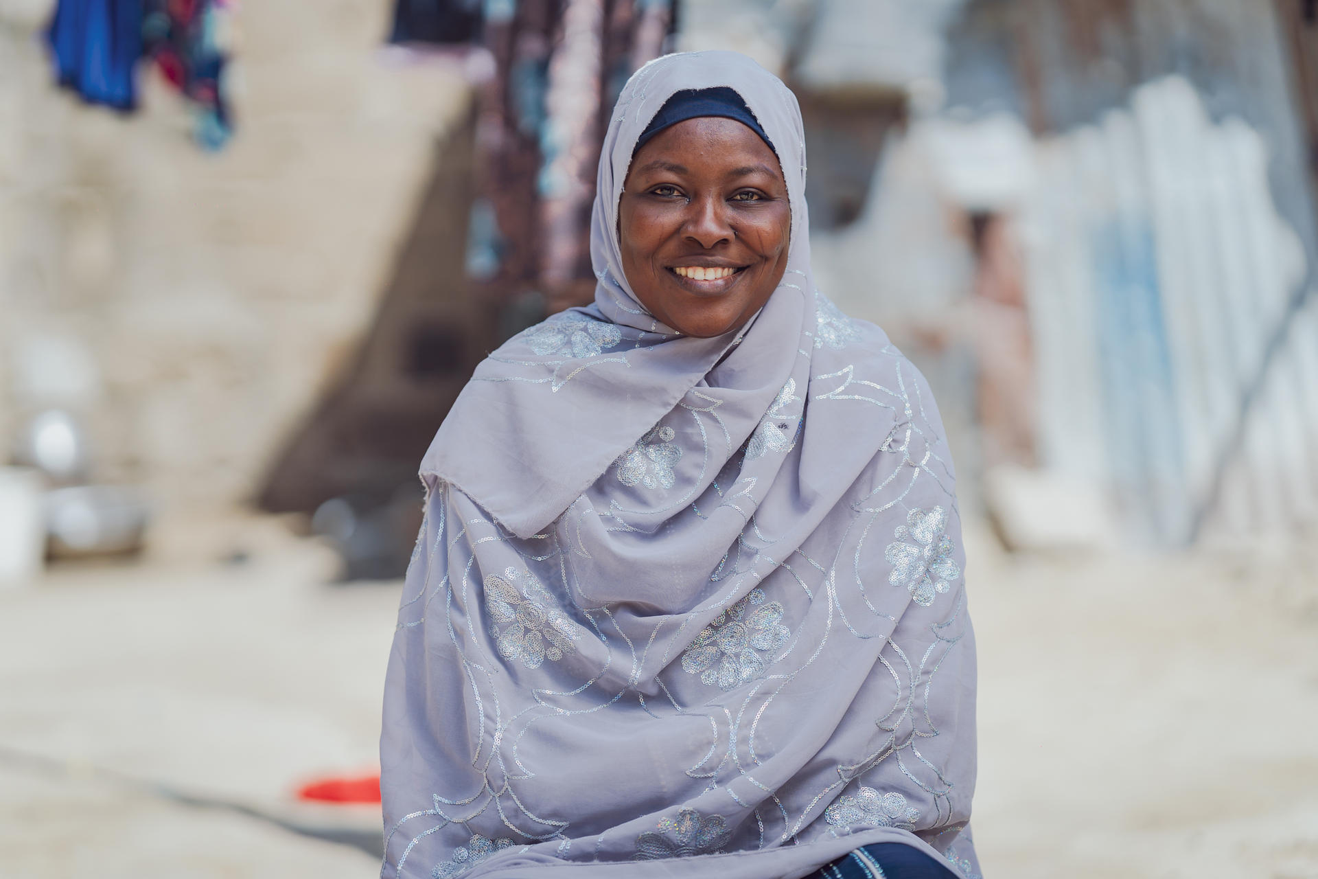A smiling woman in a grey hijab sits outdoors, with clothes hanging in the background.