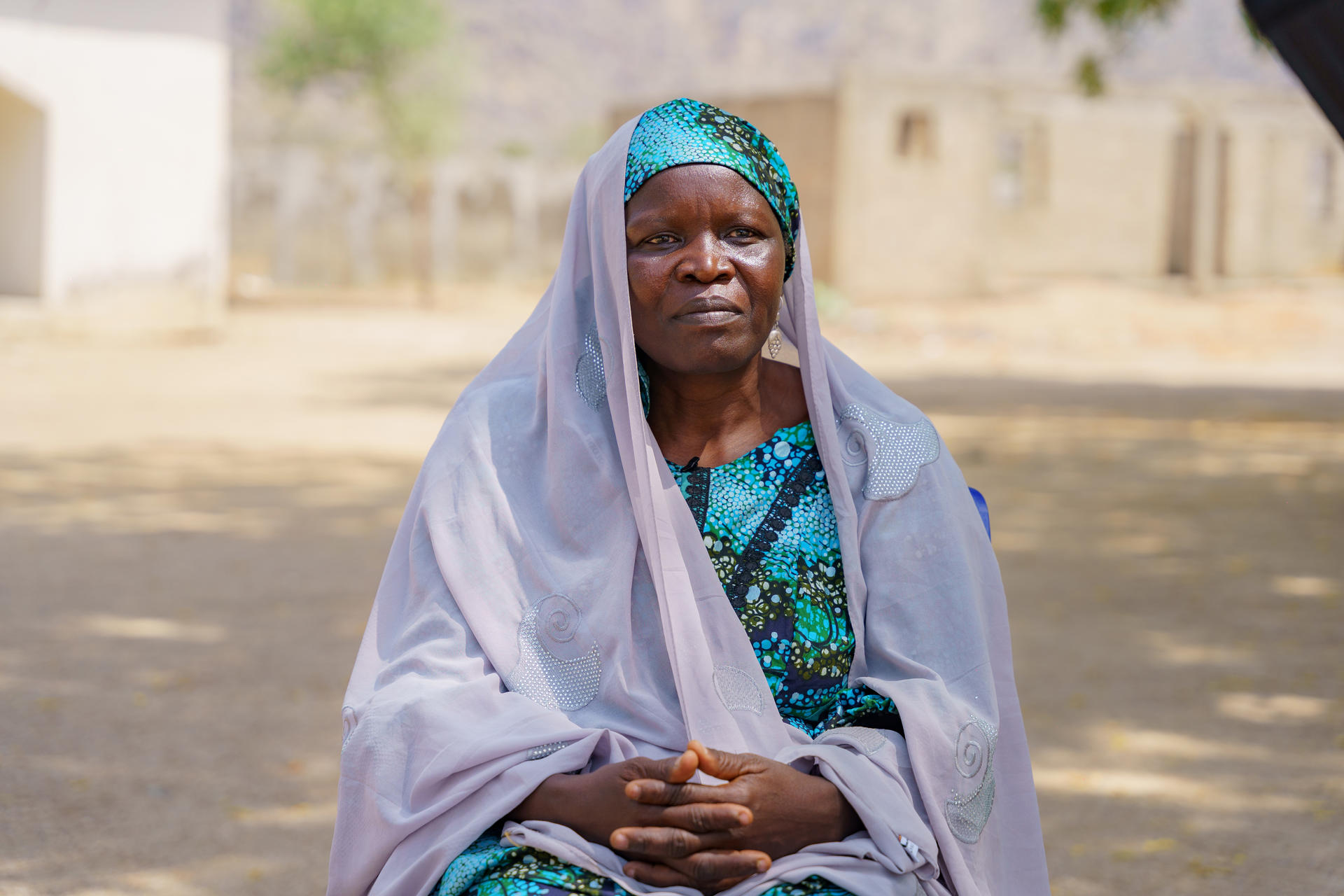 Elderly woman in a colorful outfit and headscarf, seated outdoors with a serious expression.
