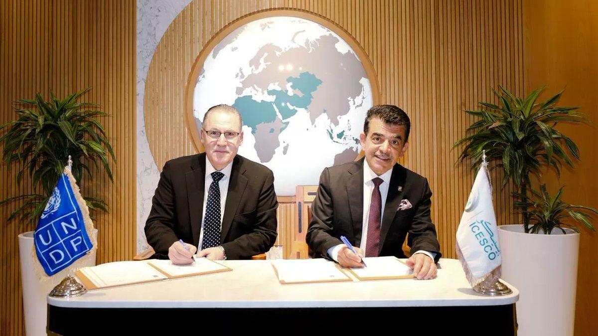 Two men in suits signing documents at a table, with plants and flags in the background.