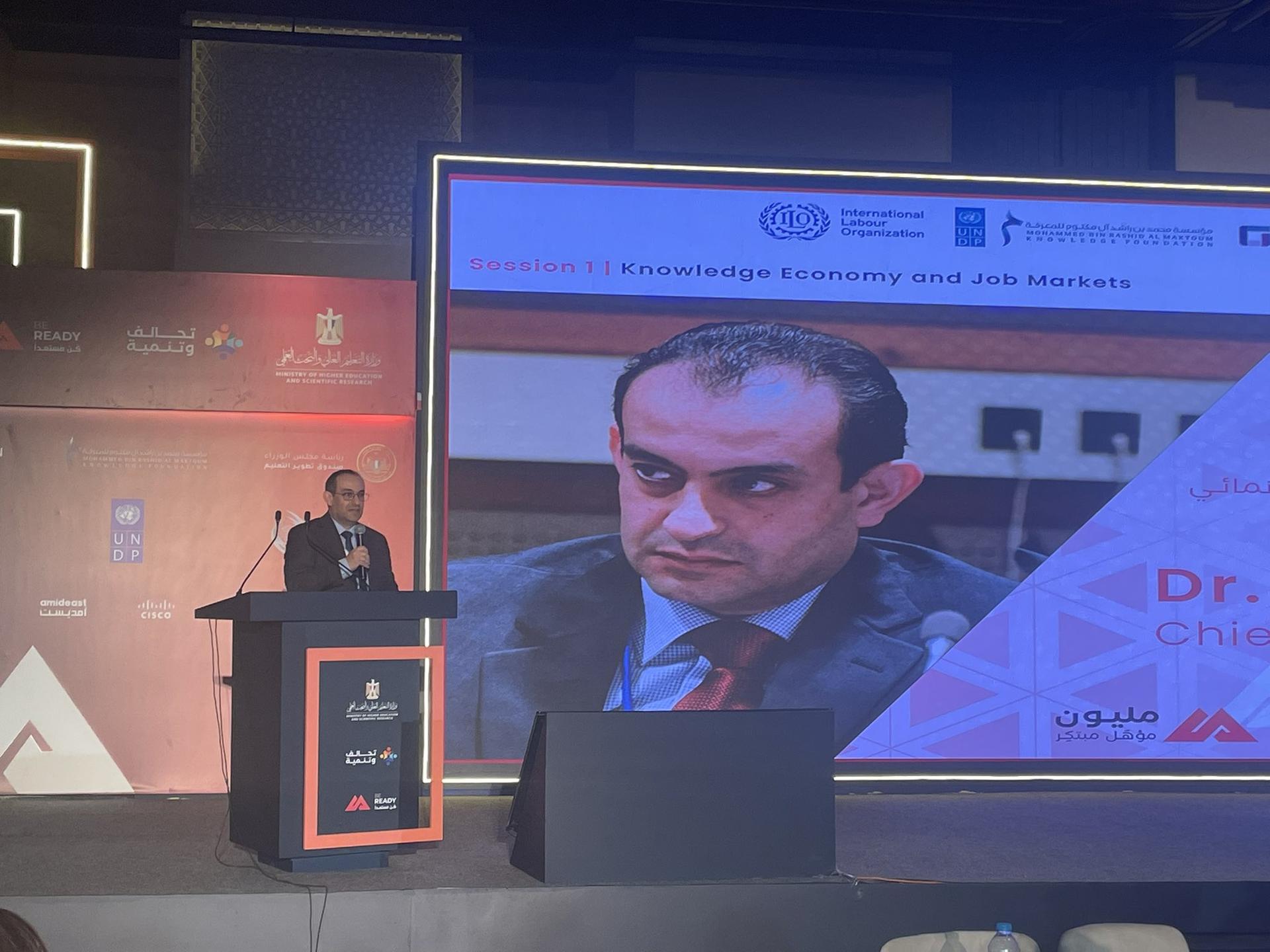 A man speaks at a podium with a projection of his image in the background.