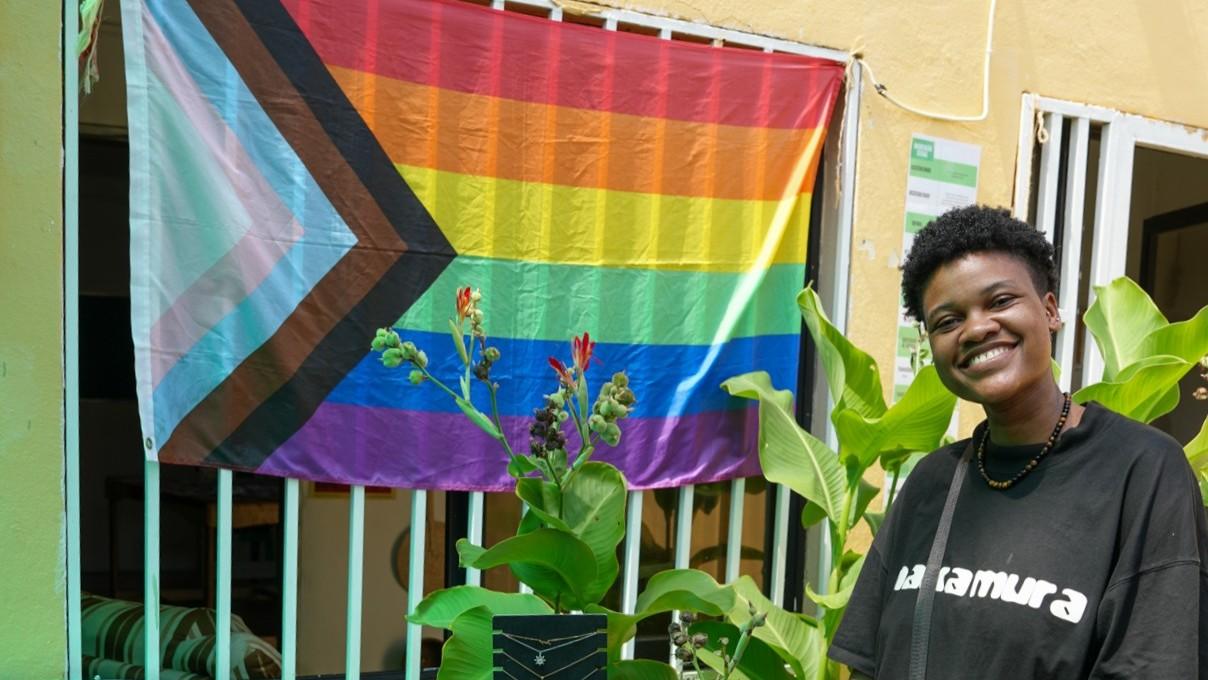 Person smiling beside a colorful pride flag and tropical plants.