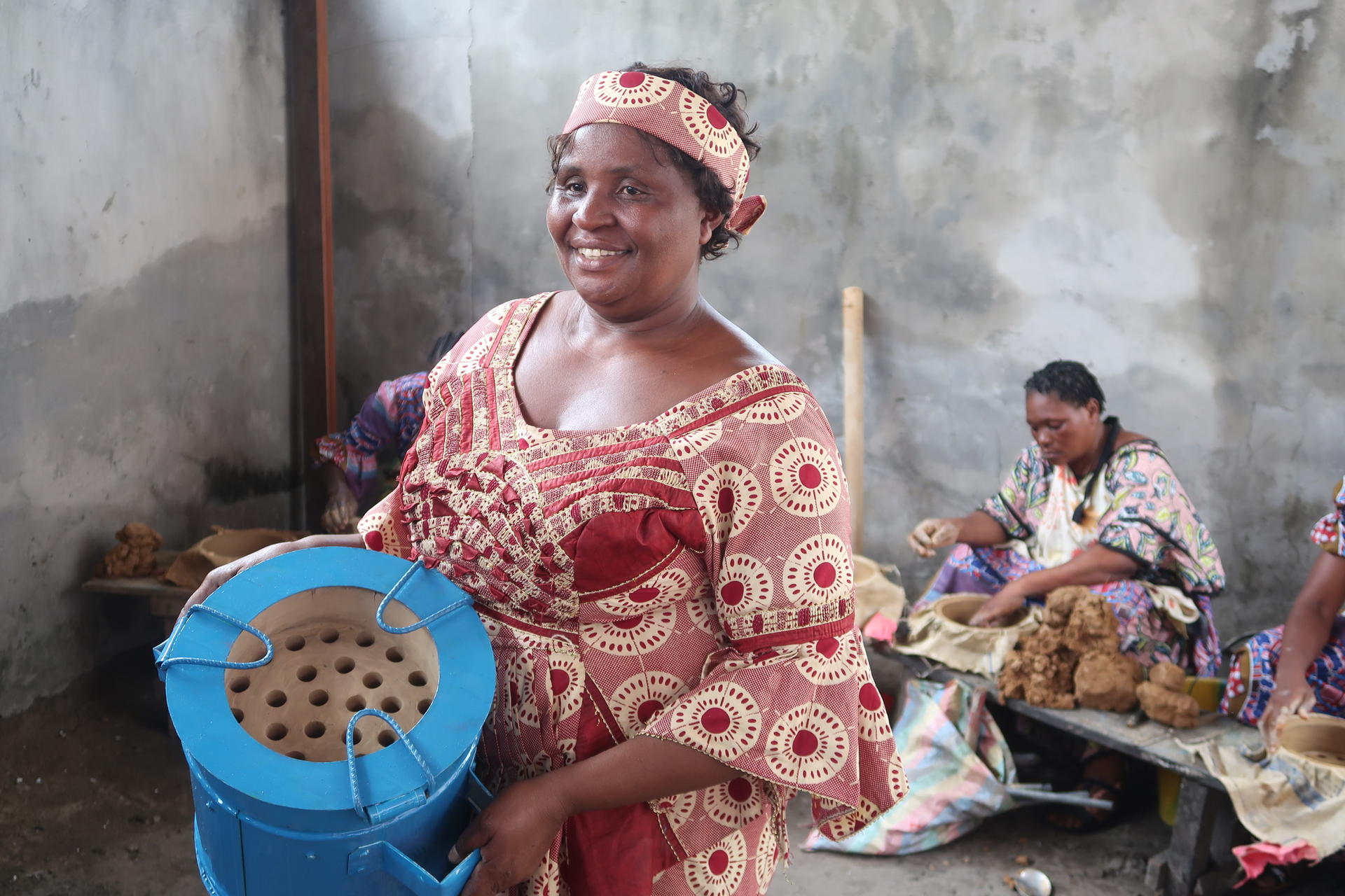 Smiling woman in a colorful dress holds a blue container, with others working in the background.
