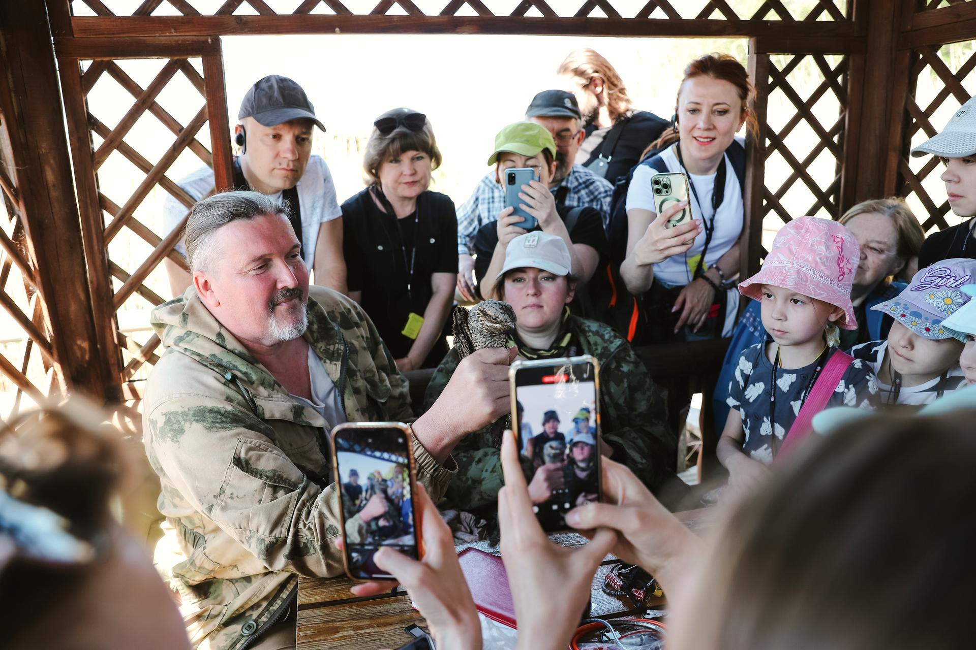 A group of children and adults gather around a man in camouflage, who is showing something in his hand.