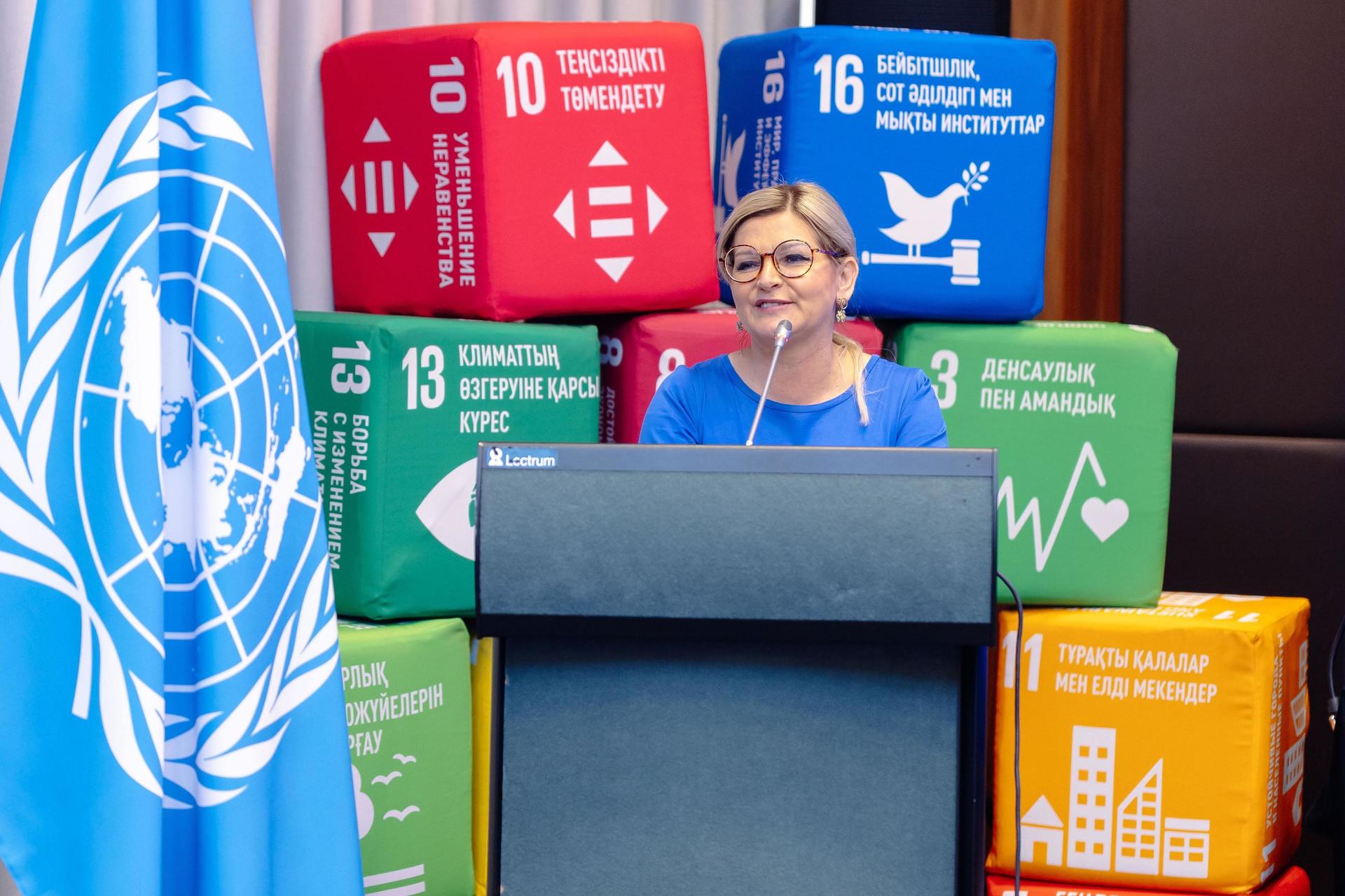 A woman stands at a podium, speaking with colorful UN SDG blocks in the background.