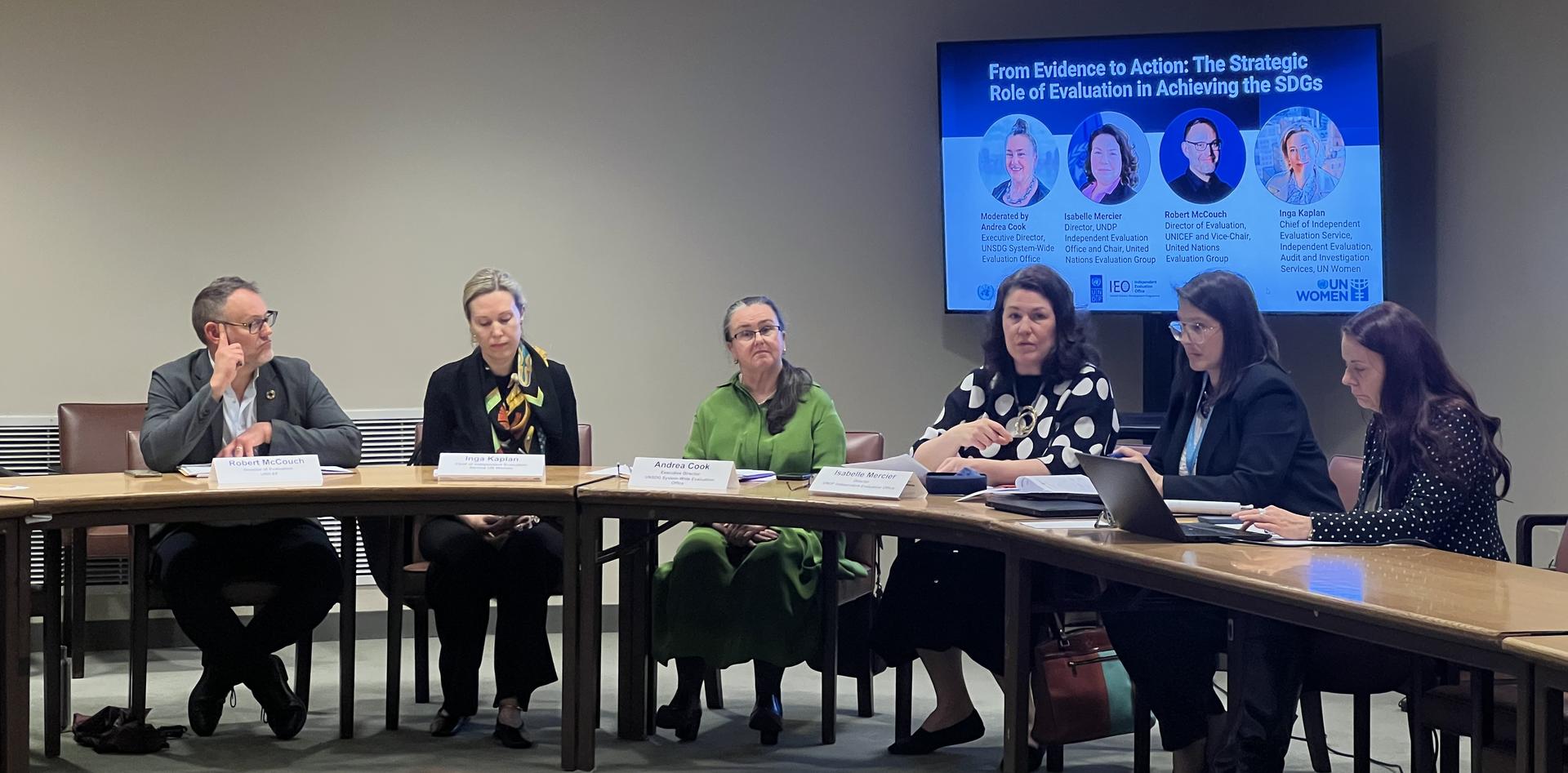 Panel discussion with five individuals seated at a table, facing an audience and a screen behind.