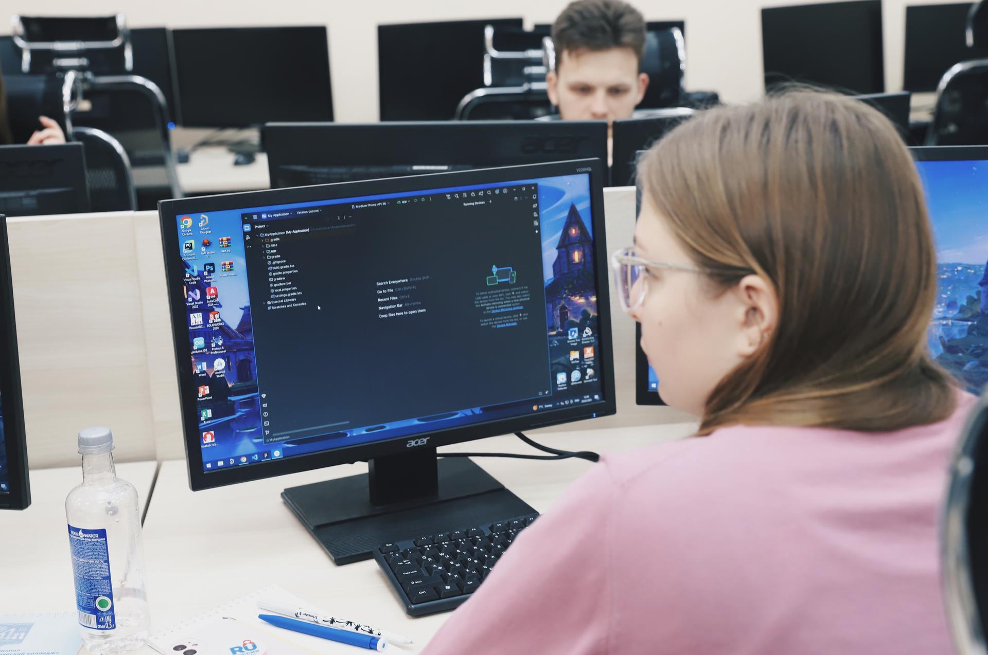 A student coding on a computer in a classroom with additional computers in the background.