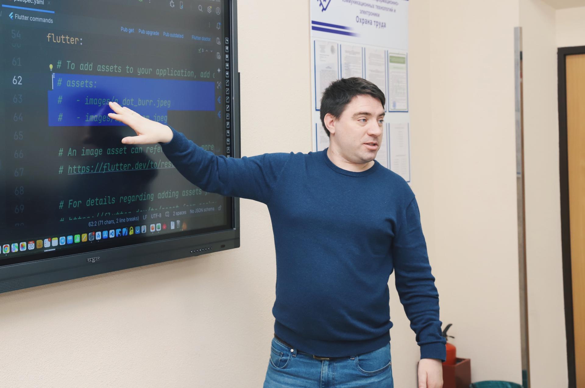 A man gestures while presenting coding information on a large screen in a classroom setting.